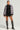 A person poses confidently wearing the MINA FAUX FUR BOMBER, featuring a patterned design with faux leather trim, paired with a black top adorned with silver accents, a black mini skirt embellished with lace details, and knee-high glittery boots. The oversized silhouette of the jacket steals the spotlight against the plain, neutral background.