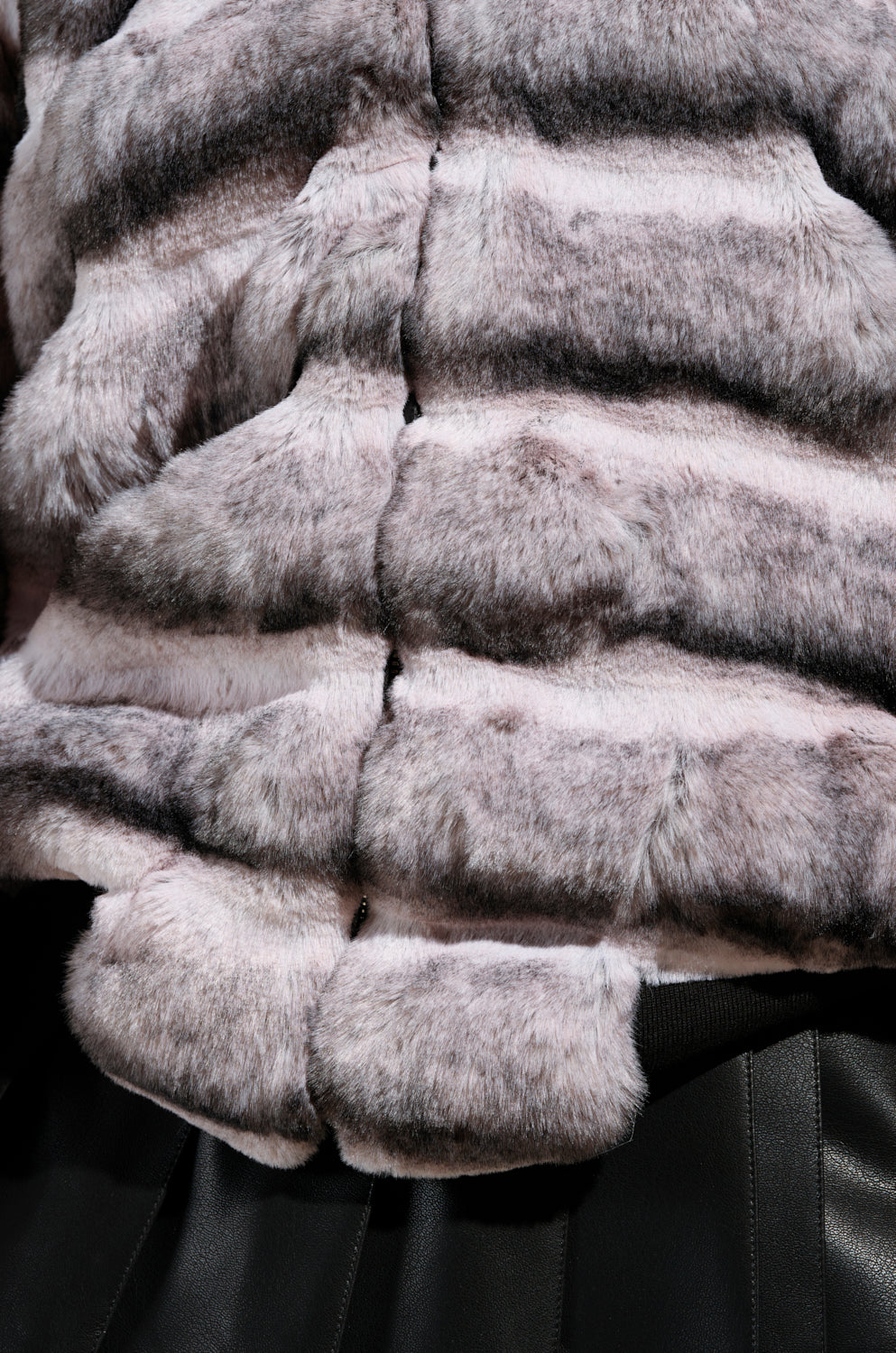 Close-up of the MINA FAUX FUR BOMBER, showcasing its gray and pink striped design with a textured, fluffy appearance and an oversized silhouette. The horizontal pattern gives it a luxurious and soft look. At the bottom of the image, part of a black garment with faux leather trim is visible.