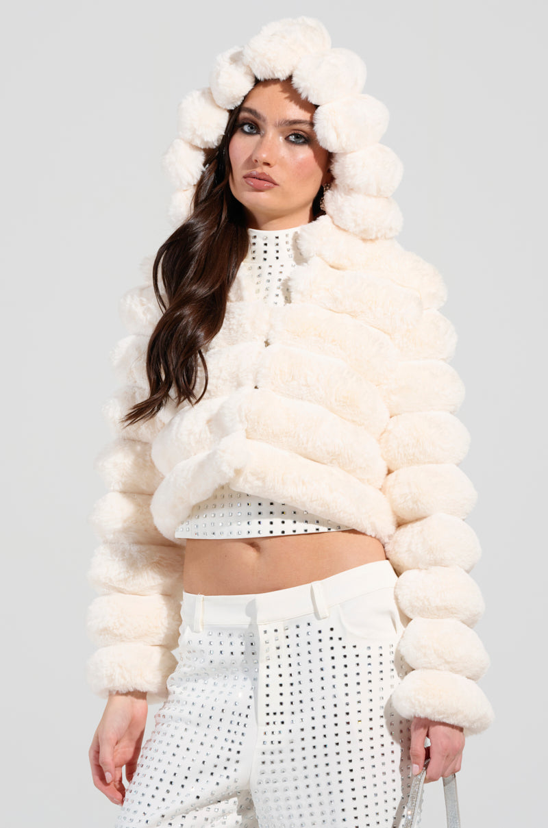A woman in a BUNNY CROPPED FAUX FUR COAT, cream-colored with fluffy texture and circular patterns, poses confidently. She pairs it with matching high-waisted pants featuring a perforated design, standing in front of a plain background.