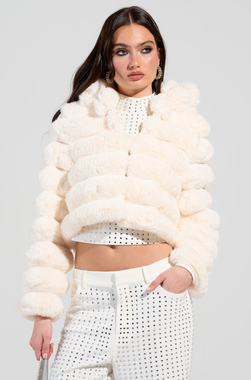 Dressed in a luxurious BUNNY CROPPED FAUX FUR COAT, a woman layers it over a white top detailed with delicate black polka dots. She completes her look with coordinating white pants featuring the same stylish pattern. Gazing thoughtfully to the side, she perfectly captures the essence of winter elegance.