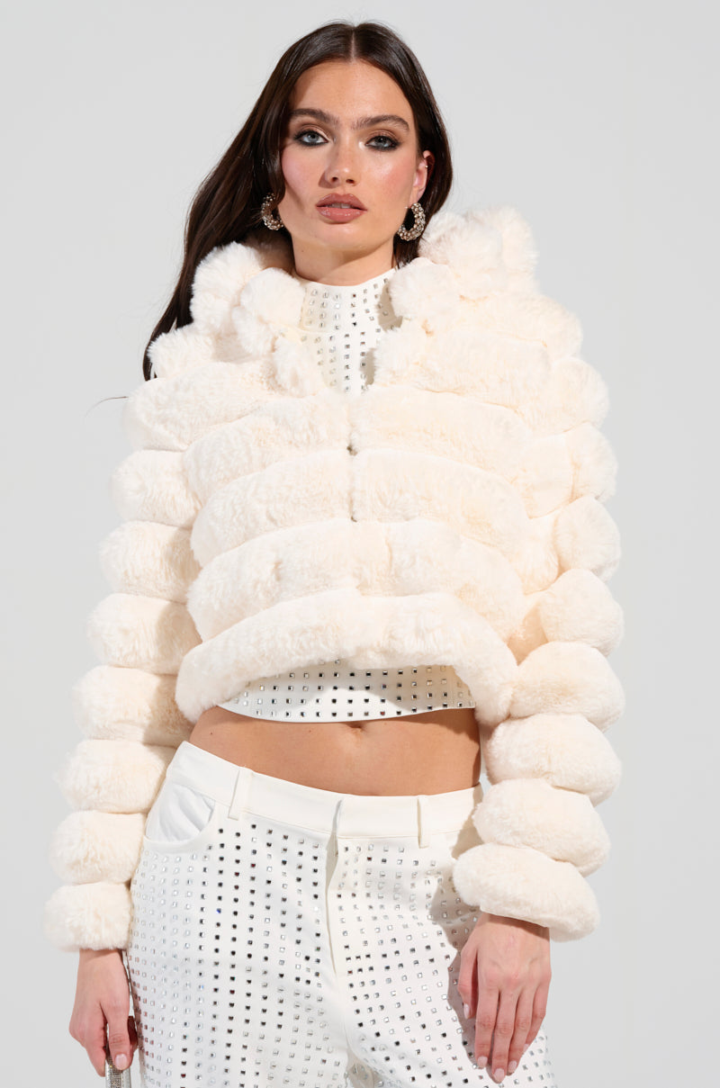 A woman dressed in a white, plush Bunny Cropped Faux Fur Coat over a black and white polka dot ensemble poses with her long, dark hair cascading down, striking a pose with one hand on her hip against a light-toned backdrop.