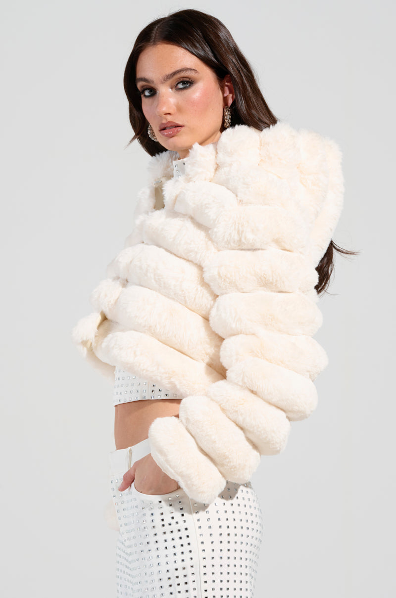 A person with long dark hair is wearing a fluffy, tiered white BUNNY CROPPED FAUX FUR COAT that evokes the charm of a Winter Wonderland Coat. They are also dressed in a white outfit adorned with small silver studs, set against a plain, light-colored background.
