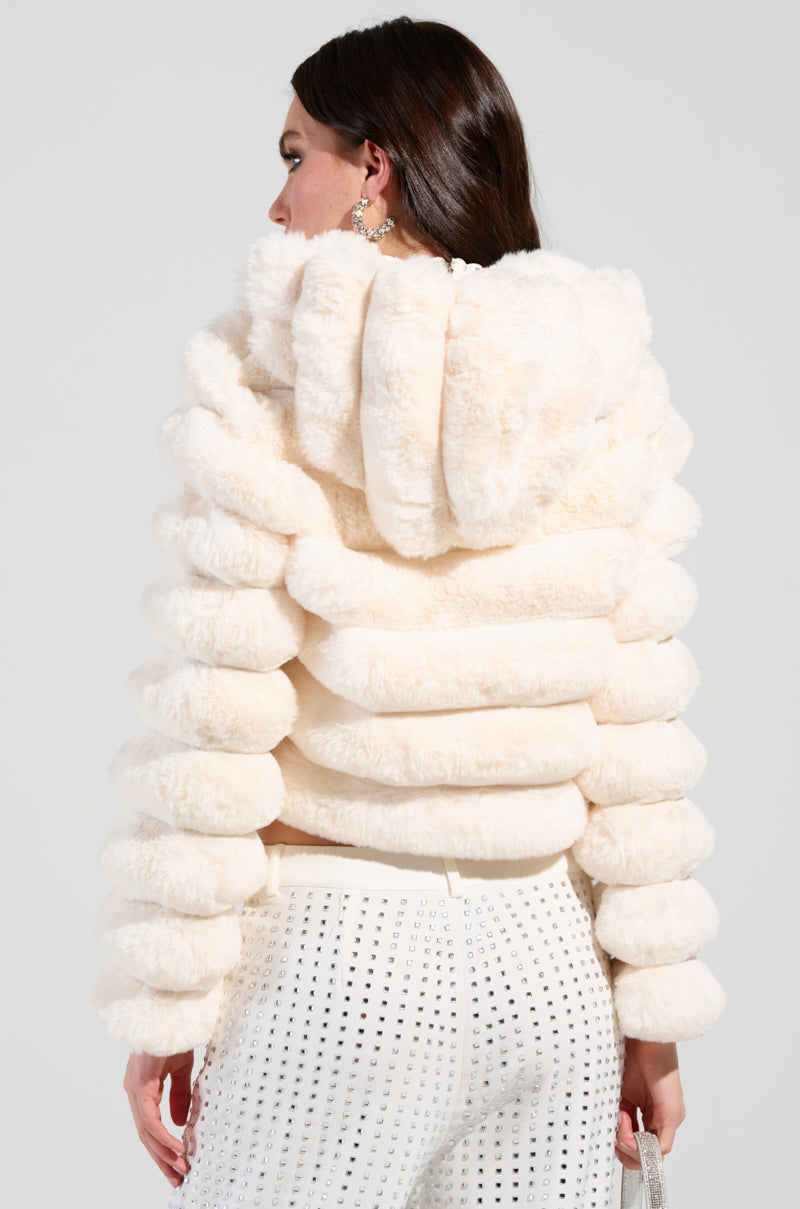 A person with long brown hair is wearing a cozy, white Bunny Cropped Faux Fur Coat with a hood, holding a silver purse. They are also dressed in white pants featuring an eyelet pattern against a plain white backdrop.