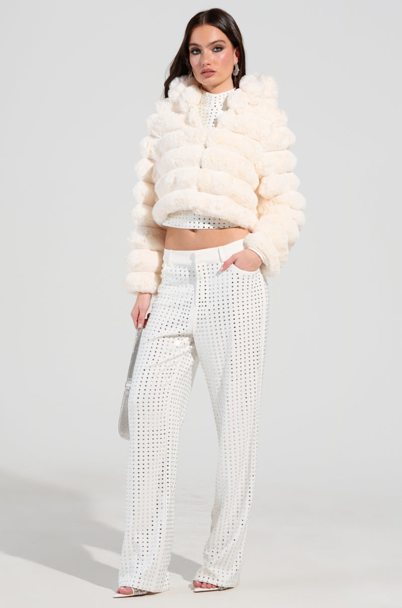 A person stands confidently, draped in a fluffy, cream-colored BUNNY CROPPED FAUX FUR COAT and white pants adorned with silver embellishments. They clutch a silver purse and wear high heels, posing against a plain background.