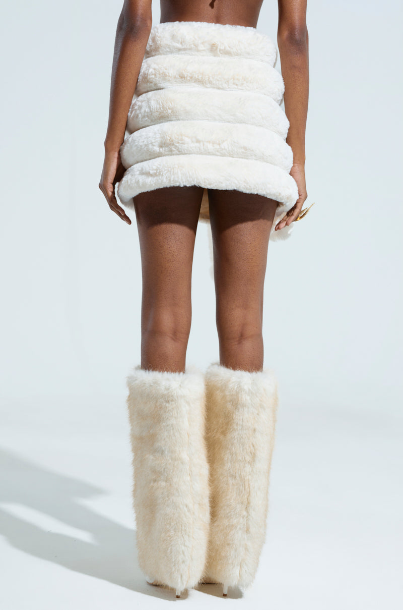A person is dressed in the BUNNY FAUX FUR MINI SKIRT, accompanied by matching high, fur-covered boots from the AKIRA Label. The fluffy, white layers are accentuated against a plain, light backdrop that emphasizes the avant-garde fashion ensemble and highlights the chic faux fur design.