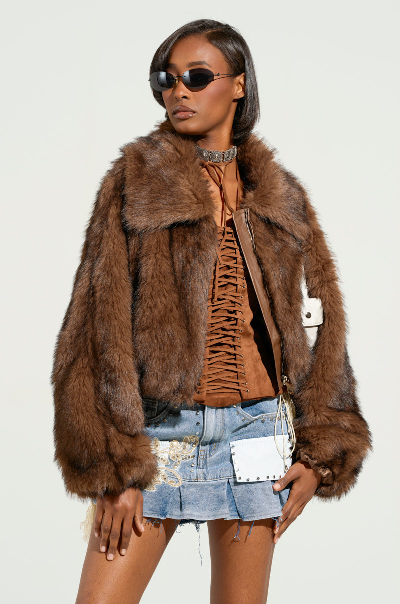 Person striking a confident pose against a simple background, wearing sunglasses and the KAI FAUX FUR BOMBER featuring metallic silver zips. The stylish crisscross top pairs perfectly with the denim skirt adorned with patches and embellishments.