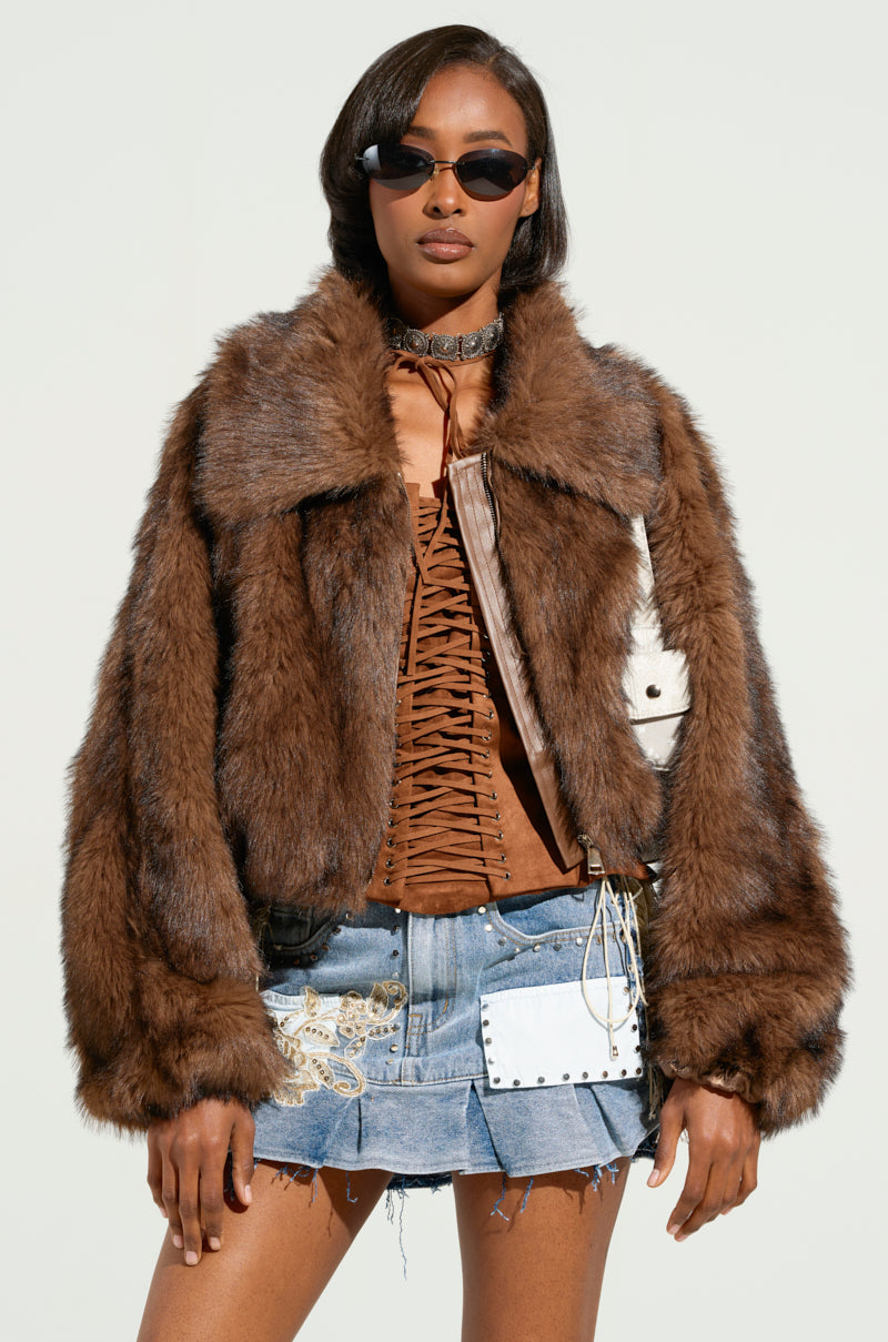 A person wearing sunglasses and the KAI FAUX FUR BOMBER featuring a metallic silver zip over a lace-up brown top, paired with a denim skirt embellished with decorative patches, stands against a light and unobtrusive background.