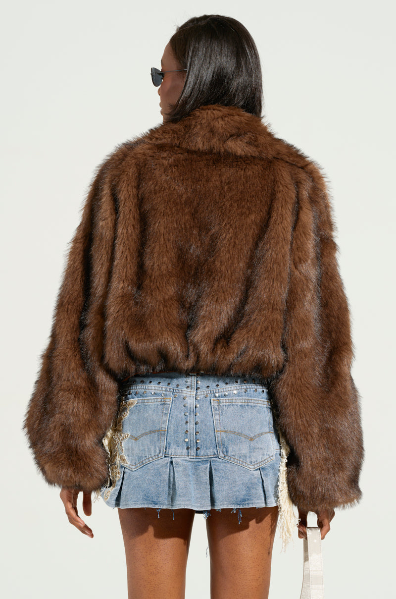 A person stands with their back to the camera, wearing the KAI FAUX FUR BOMBER and a denim mini skirt adorned with beaded details. They have long dark hair and are holding a small, light-colored handbag featuring a metallic silver zip.