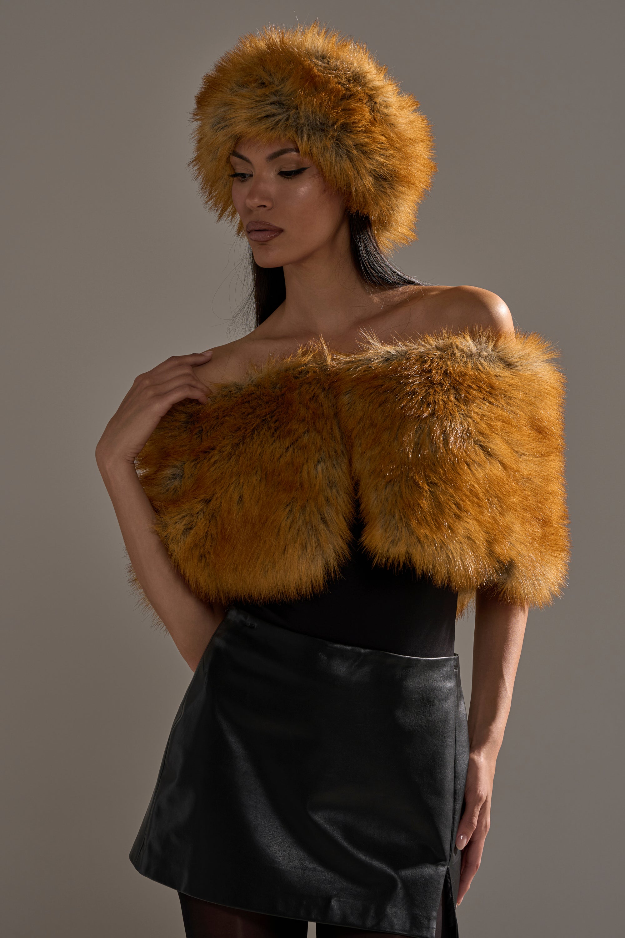 Wearing the REALLY LIKE THAT FAUX FUR SHAWL and a matching orange faux fur hat over a black top and leather skirt, a woman stands against a gray background, gently touching the soft shawl on her shoulder. 3