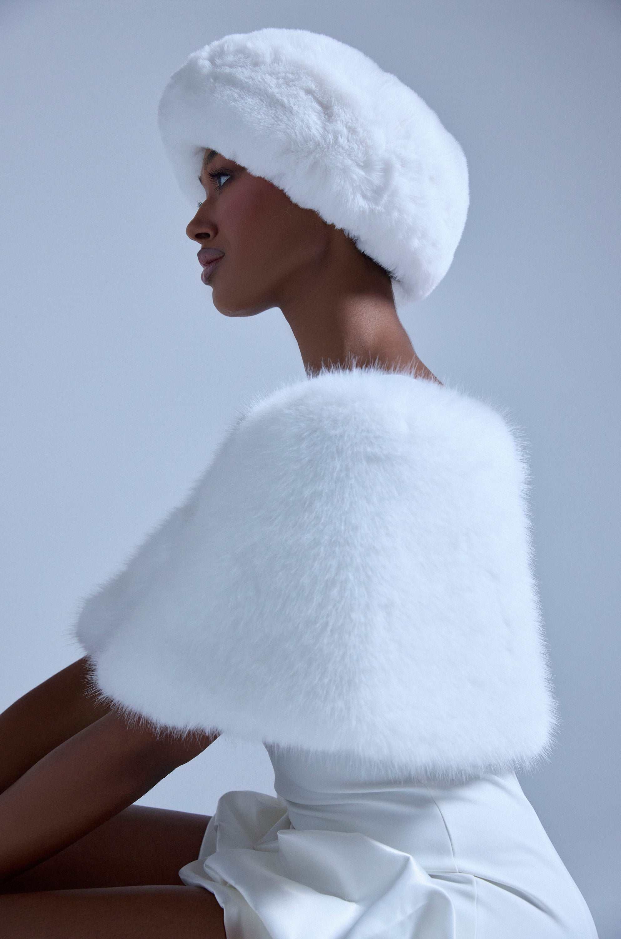 Against a light gray background, a woman in profile wears the CLASSIC FAUX FUR SHAWL and a fluffy white hat over a white dress. The elegant, winter-inspired look features soft faux fur for timeless charm. 1