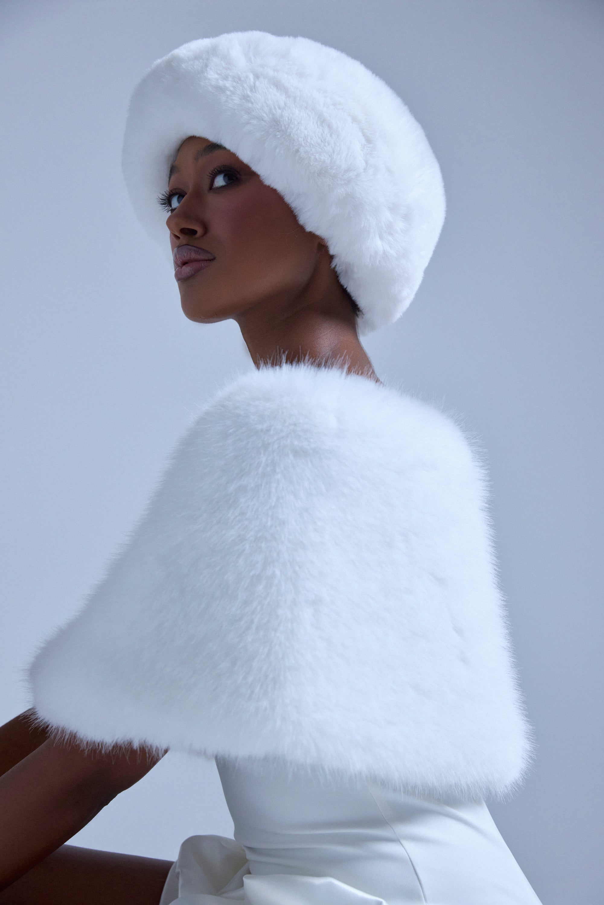 A woman wearing a white hat and the CLASSIC FAUX FUR SHAWL poses elegantly, glancing over her shoulder against a light gray background with confidence. 2