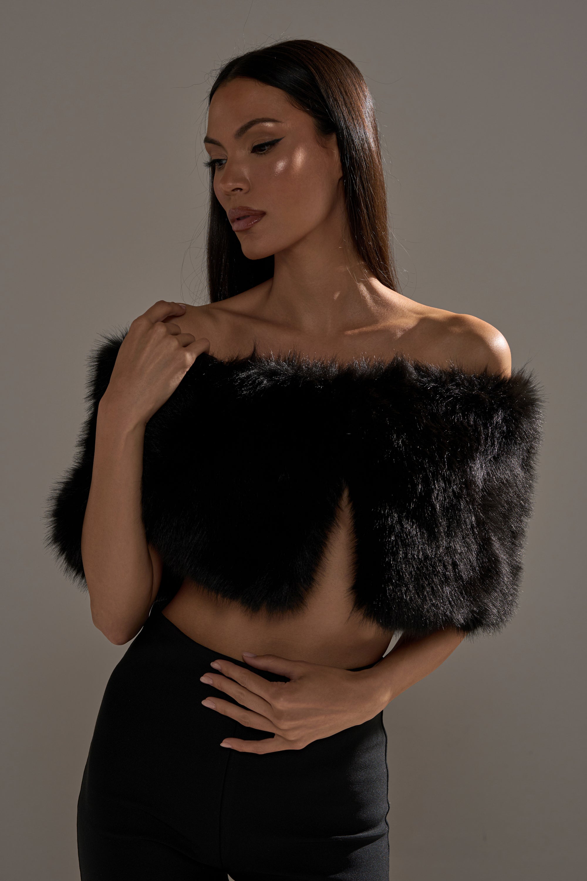 A woman with long dark hair poses against a plain background, wearing the CLASSIC FAUX FUR SHAWL draped off her shoulders and paired with black pants. Her gaze is lowered, with one hand near her chest and the other resting on her waist.