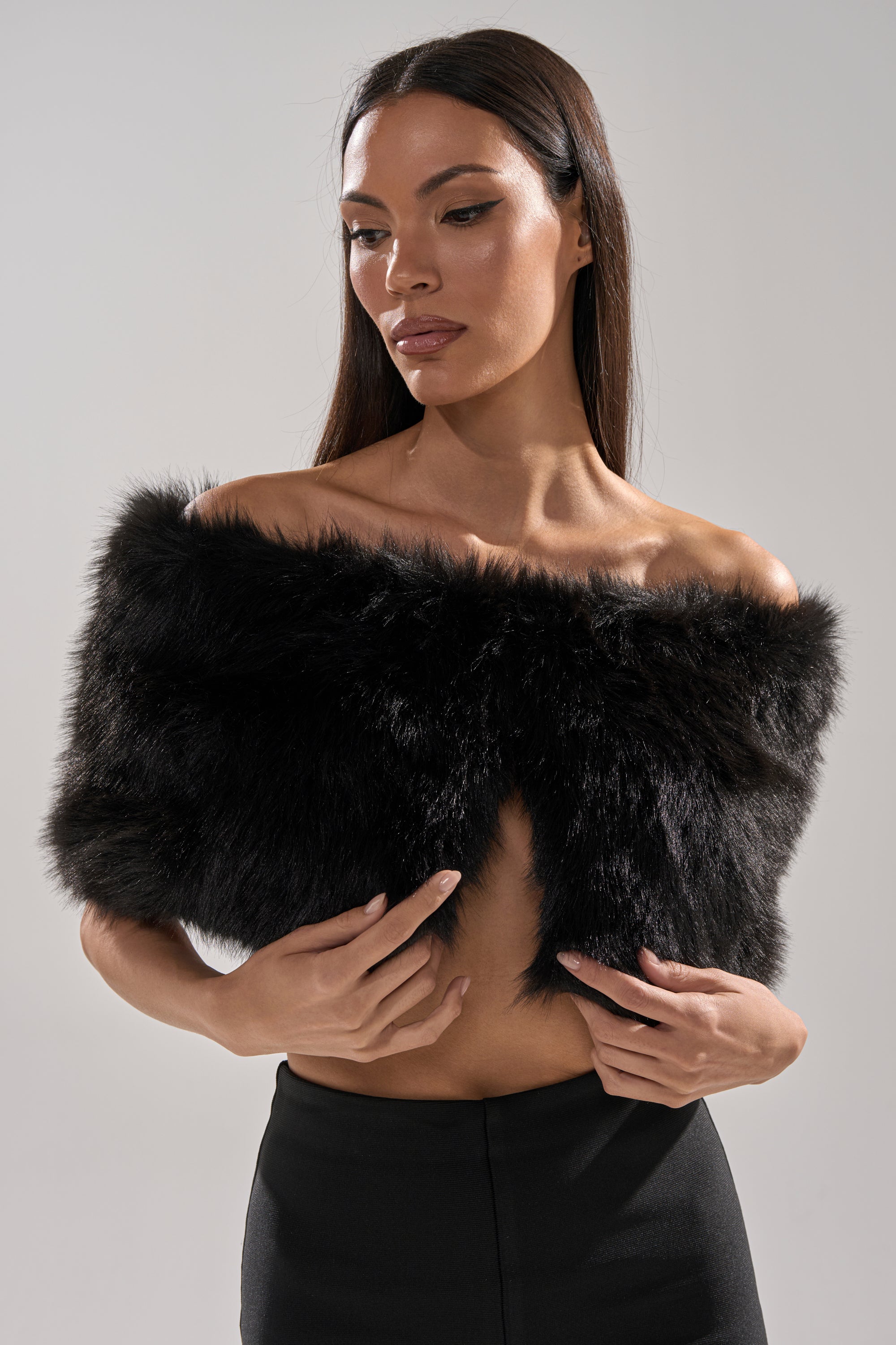 A woman with straight brown hair poses against a plain light background, looking slightly down to her left, wearing the CLASSIC FAUX FUR SHAWL in black draped off her shoulder, paired with a black skirt.