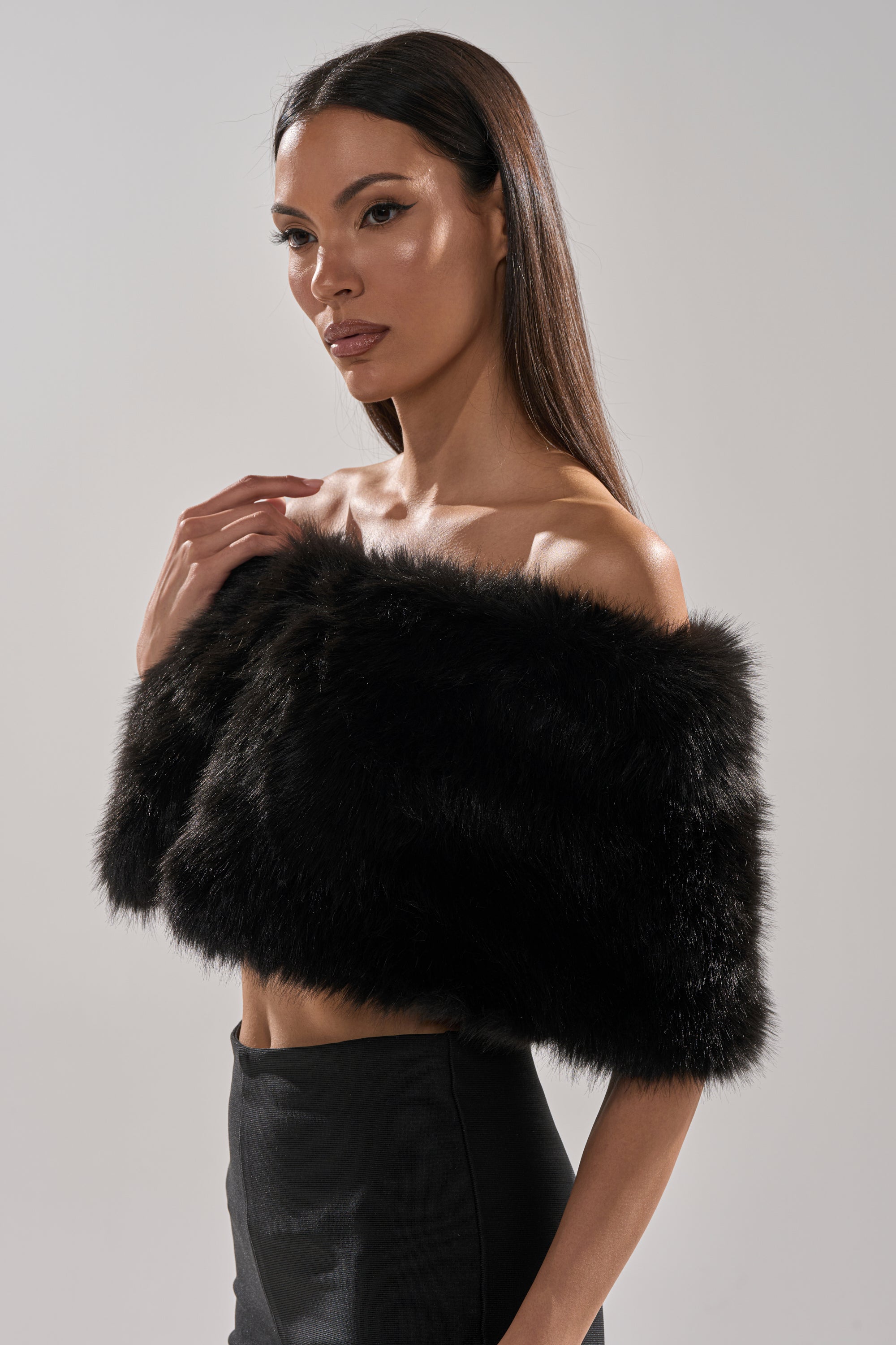 A woman with straight brown hair wears the CLASSIC FAUX FUR SHAWL, vintage-inspired with a quilted satin lining, and high-waisted black pants as she poses confidently against a plain light gray background.
