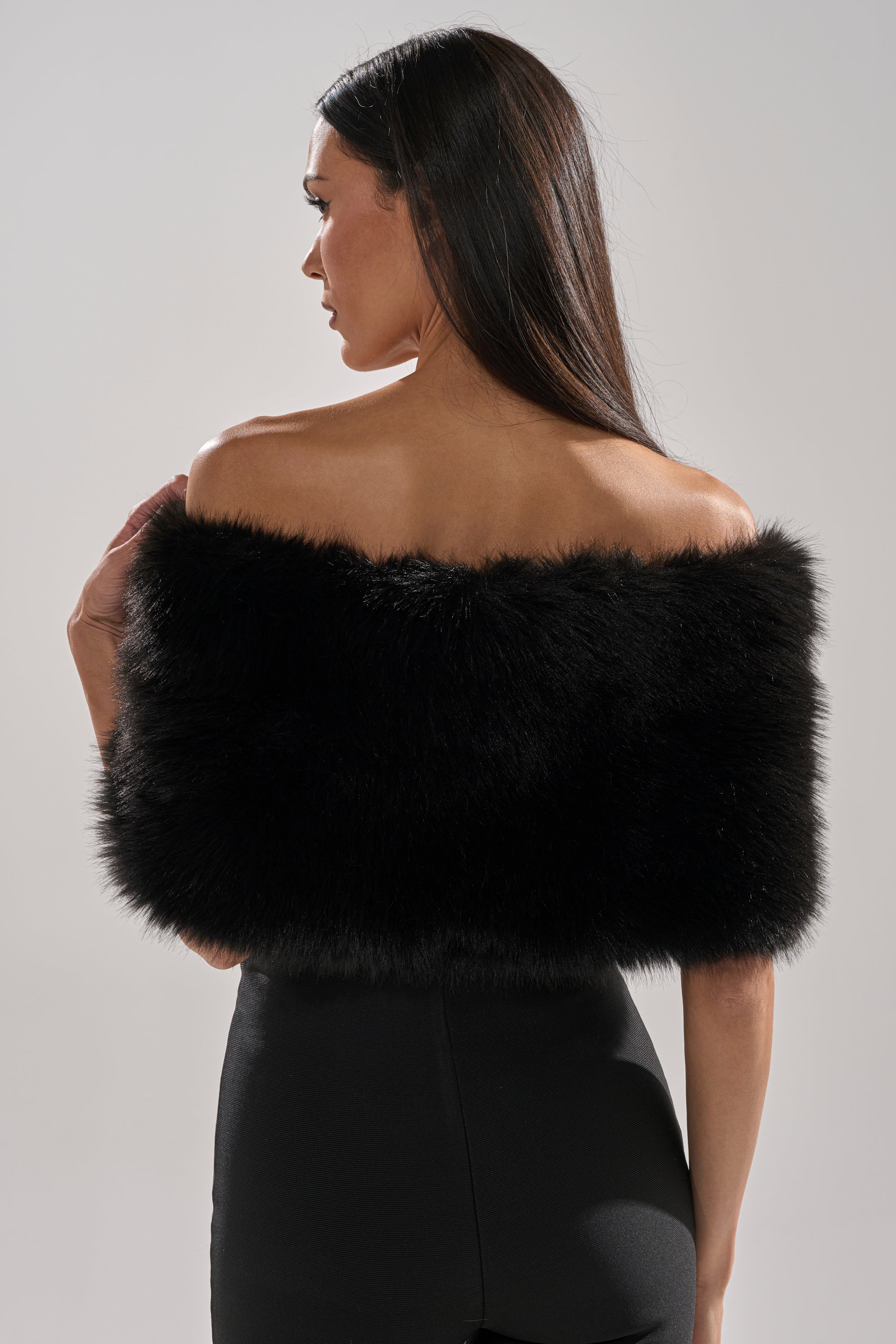 A woman with long dark hair, seen from behind, wears the CLASSIC FAUX FUR SHAWL off the shoulder with black high-waisted pants against a plain light background.