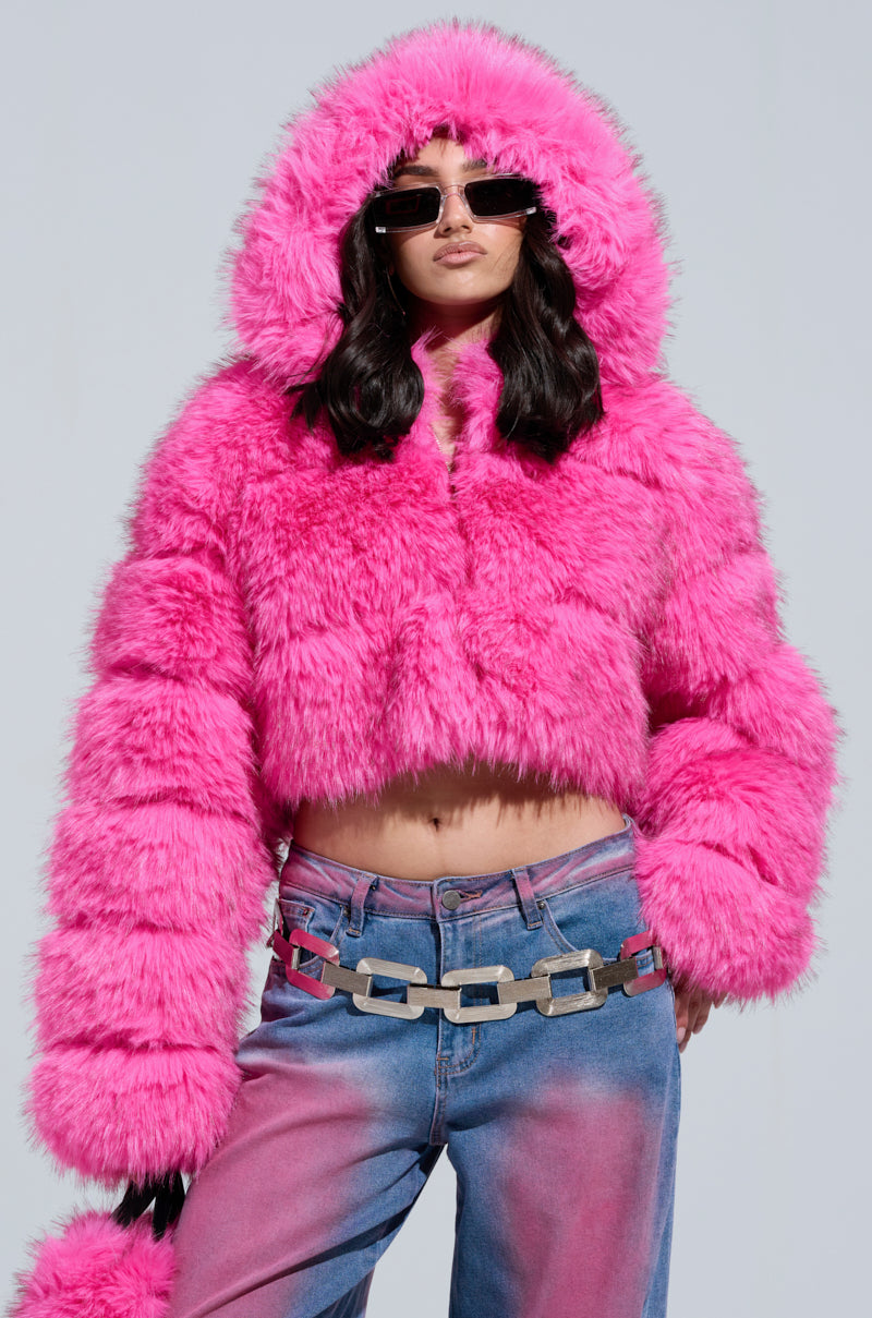 A person sports the SABRINA HOODED FAUX FUR JACKET in a vibrant pink, exuding edgy style with its cropped silhouette. Paired with pink and blue gradient jeans, dark sunglasses, and a metallic chain-link belt, they stand out against the plain gray background.