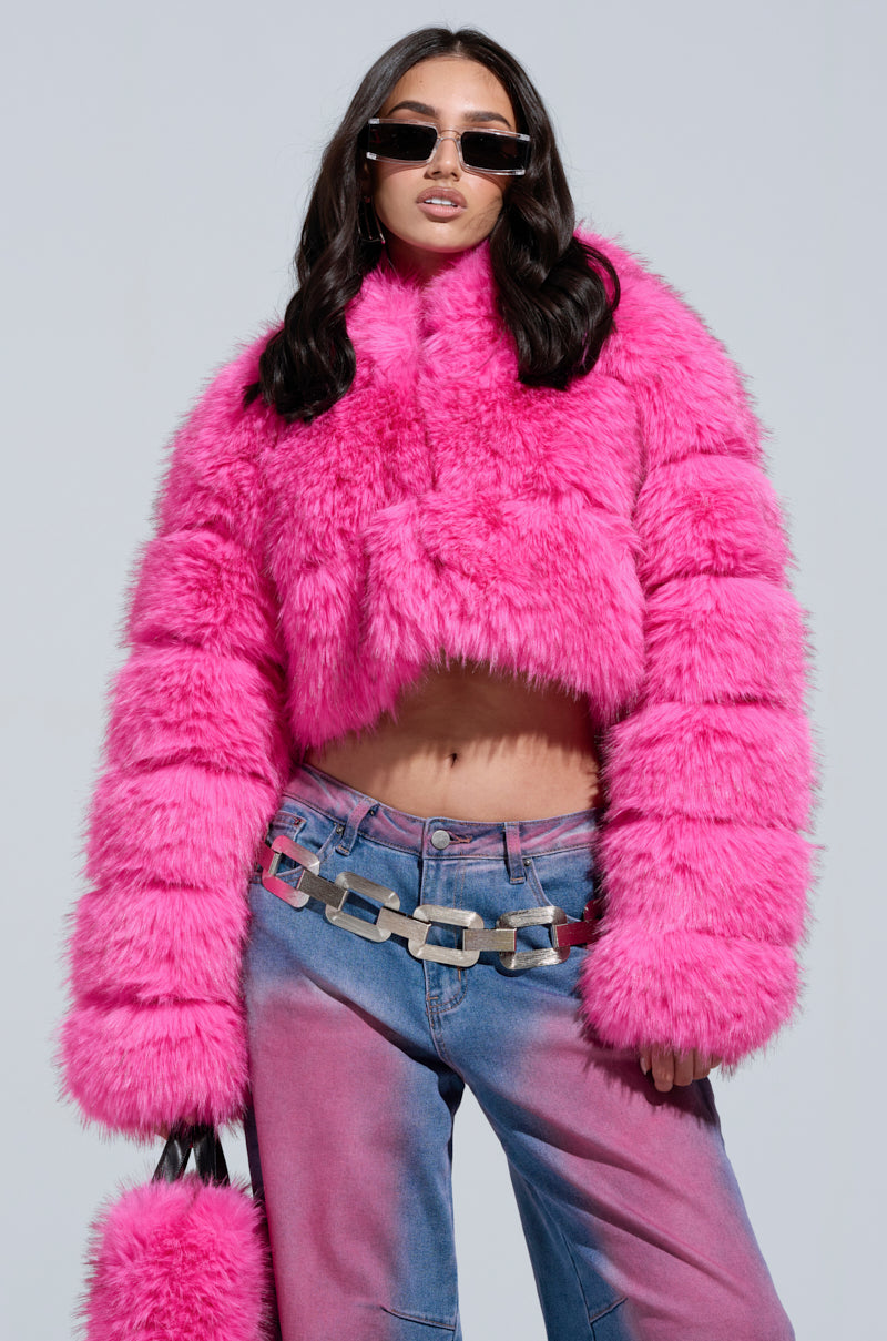 A person confidently poses wearing the SABRINA HOODED FAUX FUR JACKET in bright pink, complete with an oversized hood and sunglasses. They have long dark hair and are styled in blue jeans accompanied by a large chain belt, set against a neutral background.