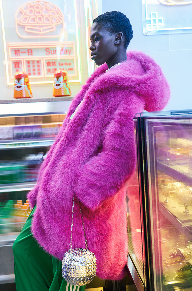A person in a vibrant DAISY FAUX FUR COAT leans against a glass display in a brightly lit store, holding a sparkly round handbag and wearing green pants. The background features colorful neon signs and various products, adding a lively touch to their stylish ensemble.