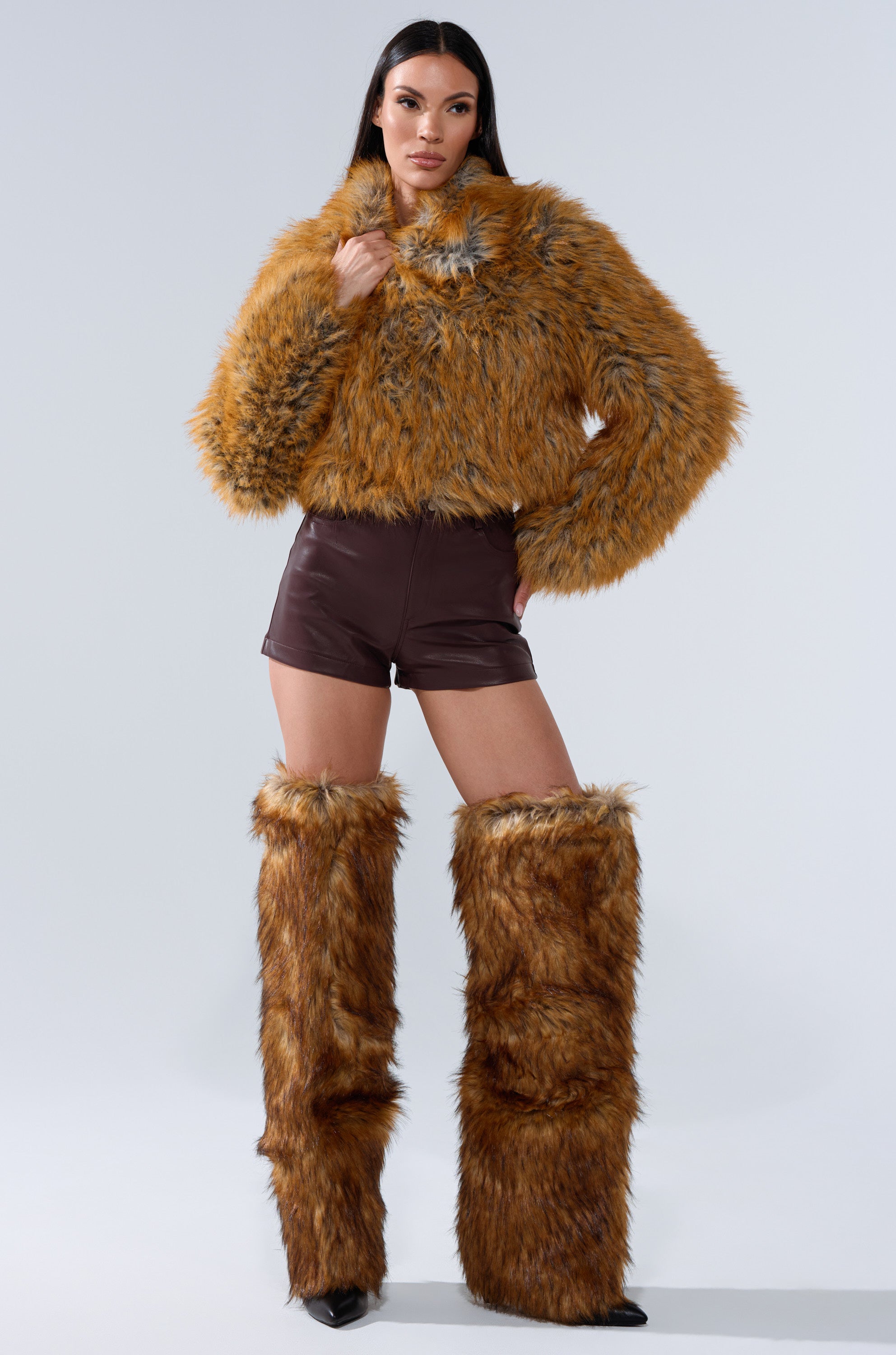 A woman poses in the LAY LOW FAUX FUR BOMBER with matching faux fur leg warmers, styled with dark brown shorts and black pointed boots against a plain light background.