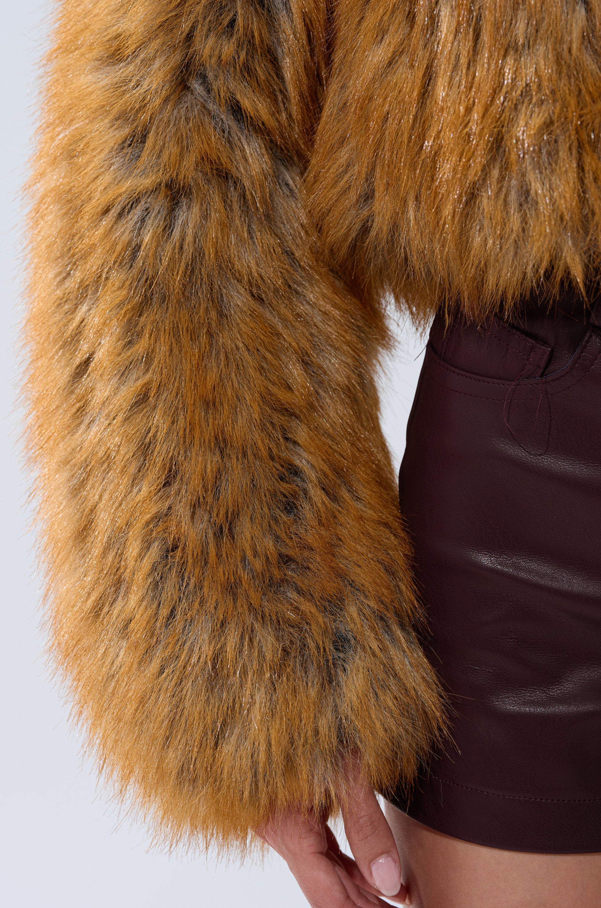 A close-up of a person wearing the LAY LOW FAUX FUR BOMBER in golden-brown with dark brown leather shorts, their hand relaxed by their side.
