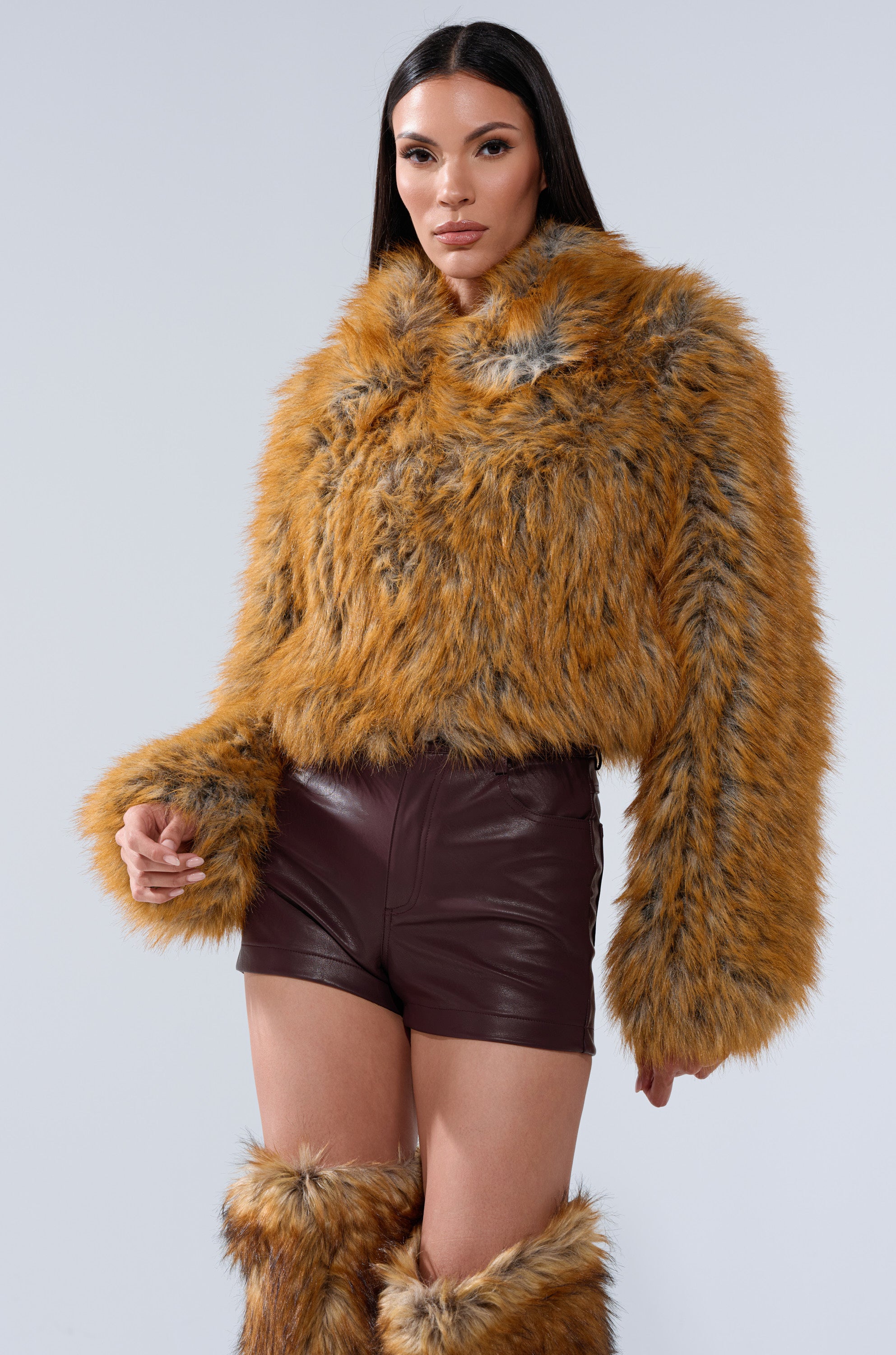 A woman with long dark hair wears the LAY LOW FAUX FUR BOMBER, matching furry leg warmers, and high-waisted dark brown leather shorts, standing against a plain light gray background.