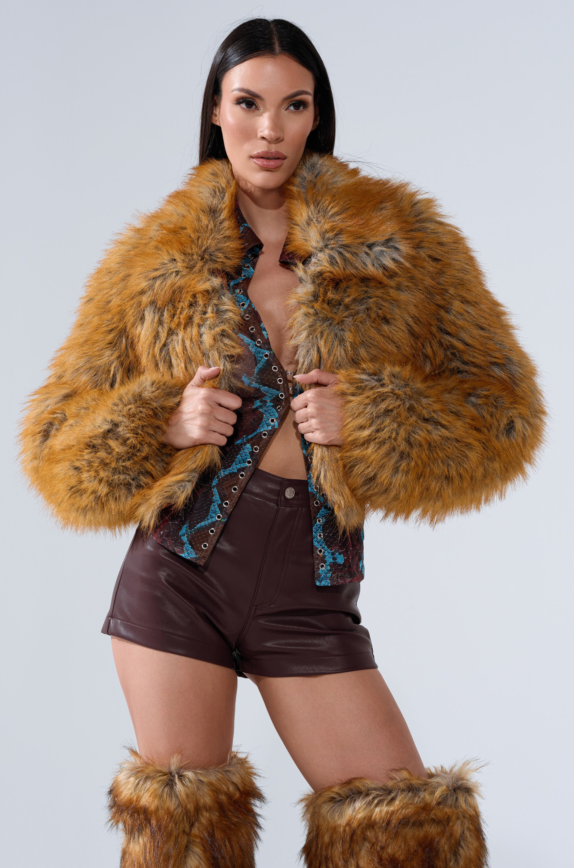 A woman poses in the AKIRA Label LAY LOW FAUX FUR BOMBER, matching orange-brown faux fur leg warmers, a patterned top, and high-waisted brown leather shorts against a plain light background.