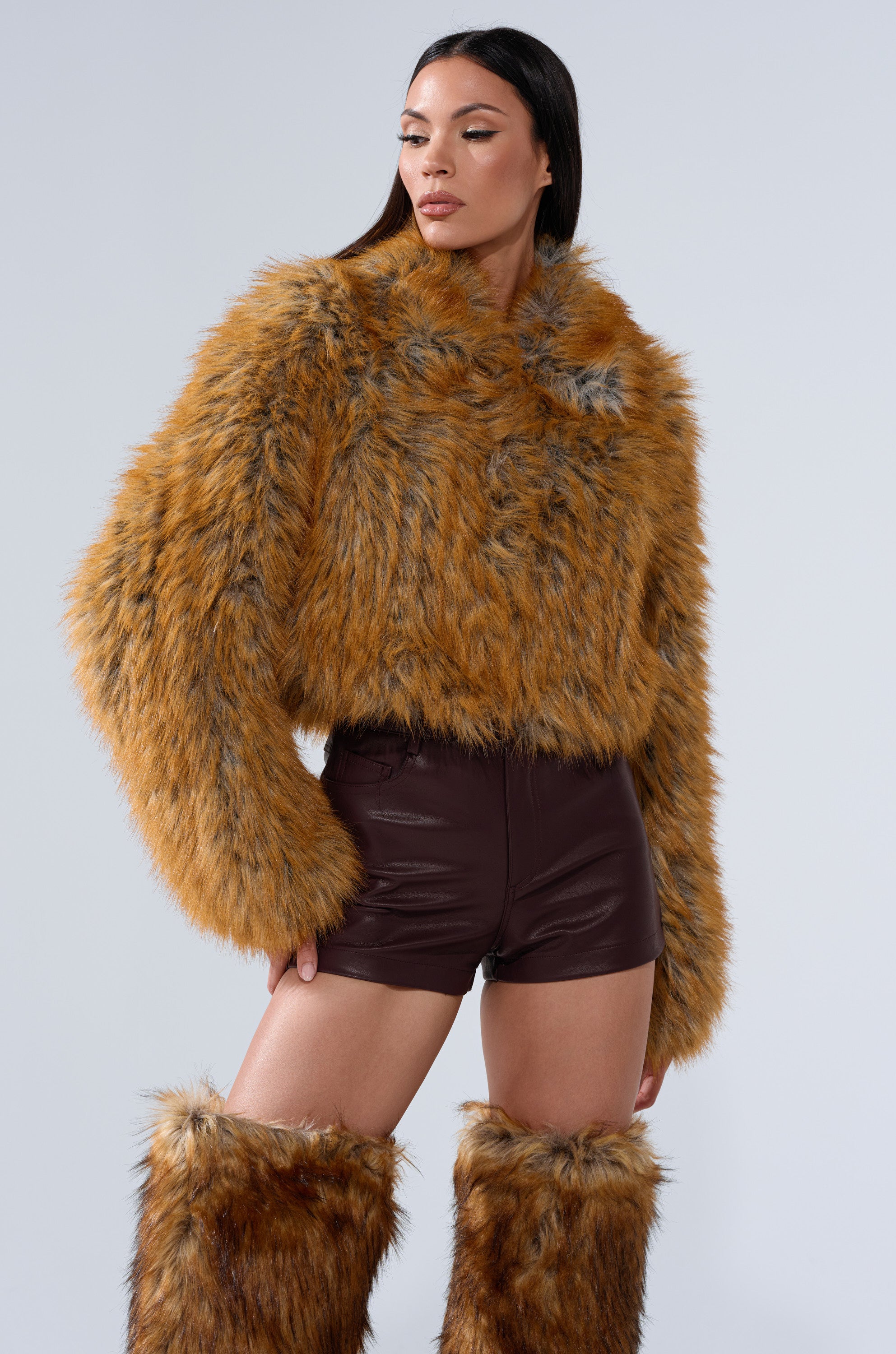 A woman poses against a light gray background wearing the LAY LOW FAUX FUR BOMBER in brown with a cropped fit, paired with matching faux fur boots and dark brown leather shorts.