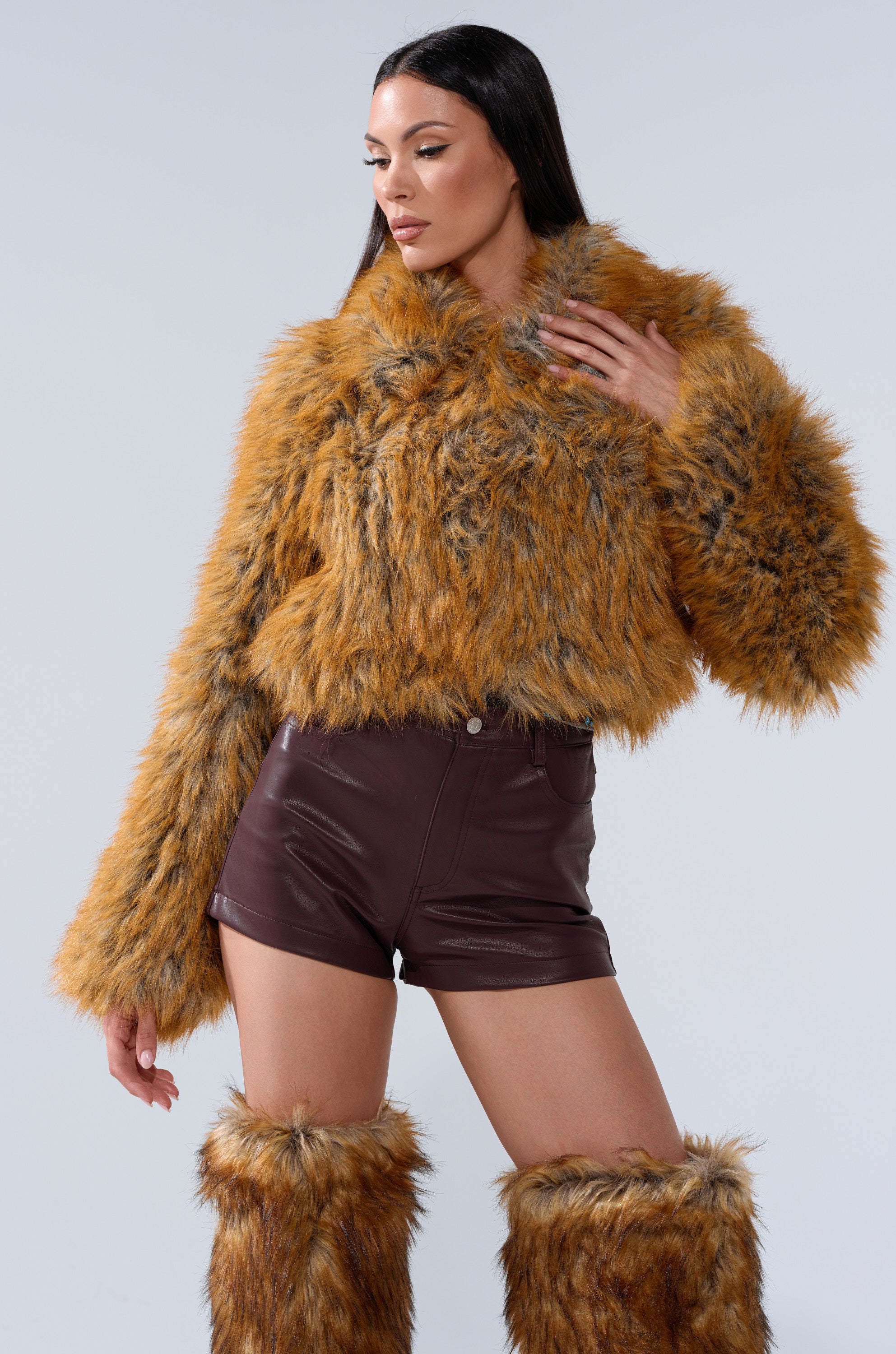 A woman models the LAY LOW FAUX FUR BOMBER with matching furry leg warmers and high-waisted brown leather shorts against a plain light background.