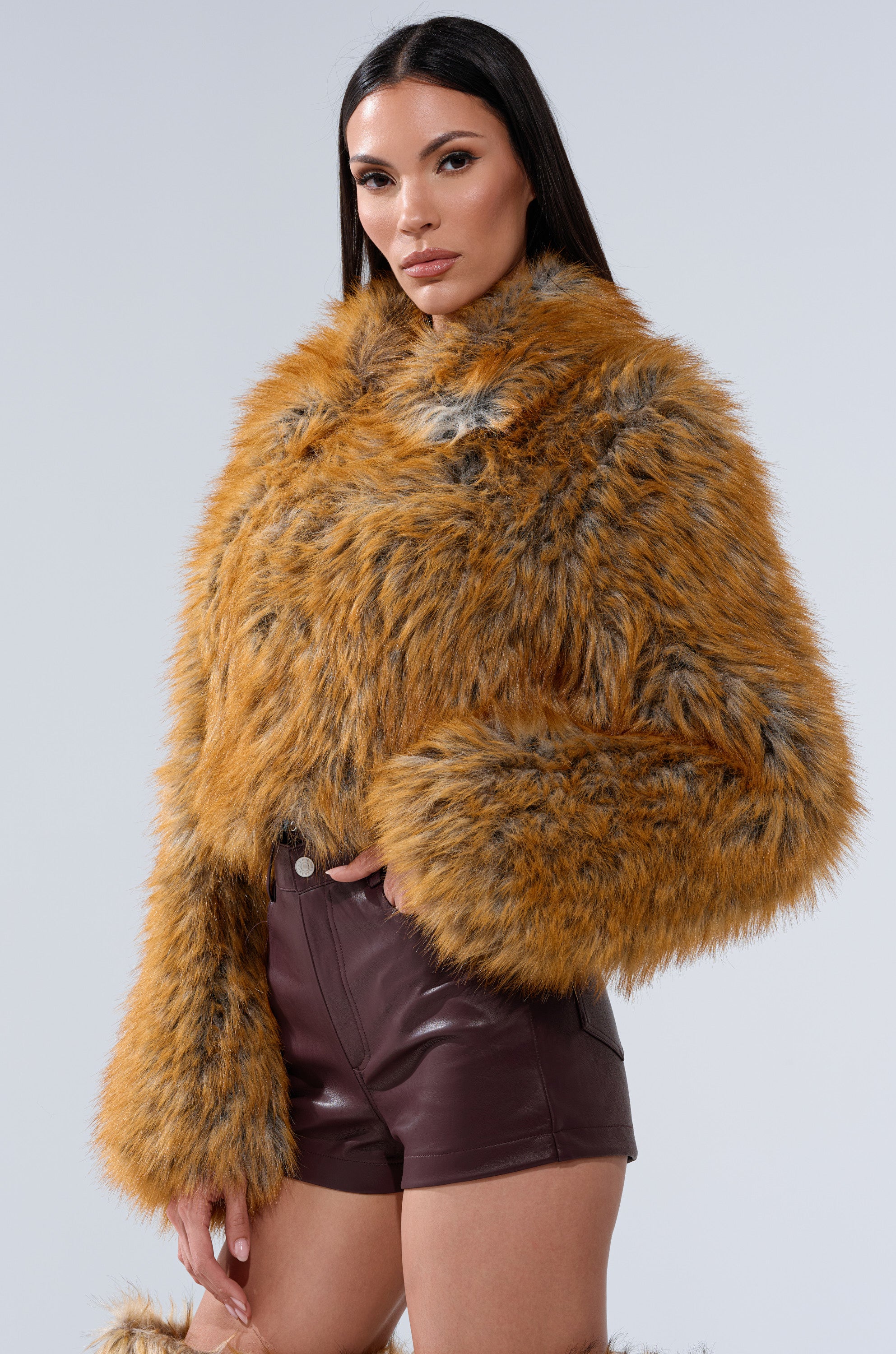 A woman with straight dark hair models the LAY LOW FAUX FUR BOMBER in orange and gray, styled with high-waisted maroon leather shorts, confidently posing against a plain light background.