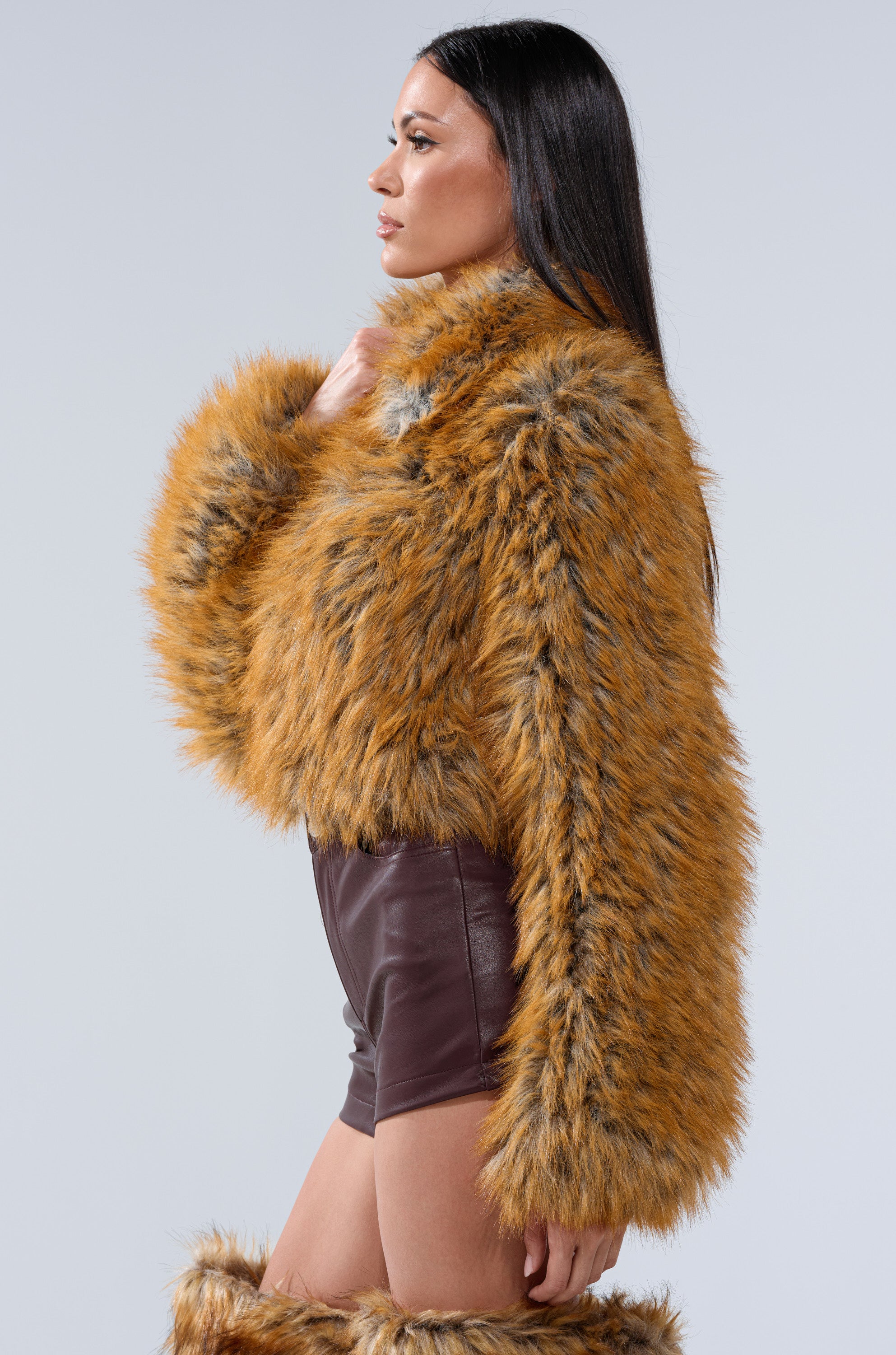 A woman with long dark hair stands in profile against a plain light gray background, wearing the LAY LOW FAUX FUR BOMBER—an orange-brown cropped faux fox fur jacket—paired with dark shorts.