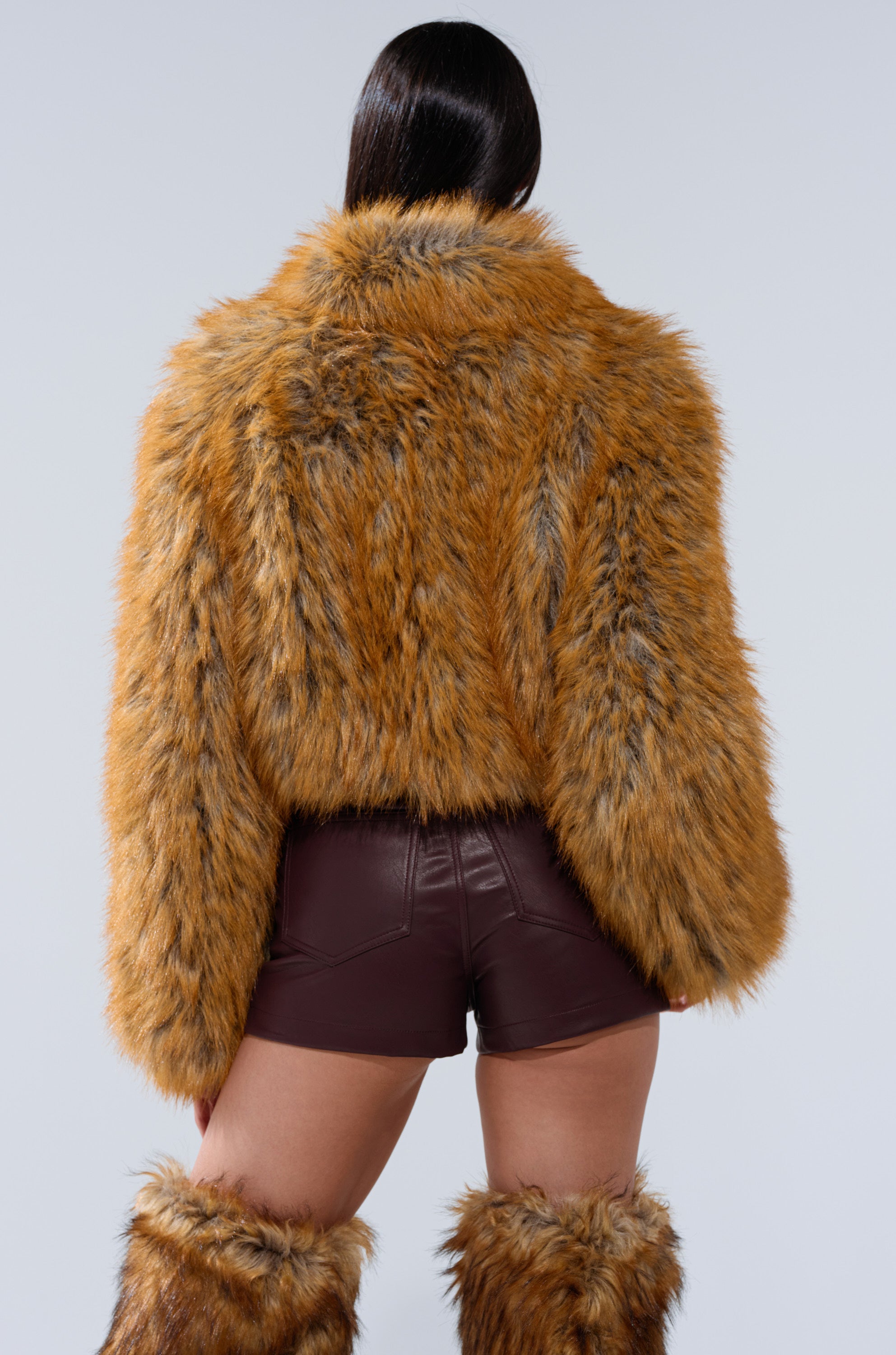 A person faces away from the camera wearing the LAY LOW FAUX FUR BOMBER in orange-brown, matching furry leg warmers, and dark brown leather shorts against a plain light gray background.