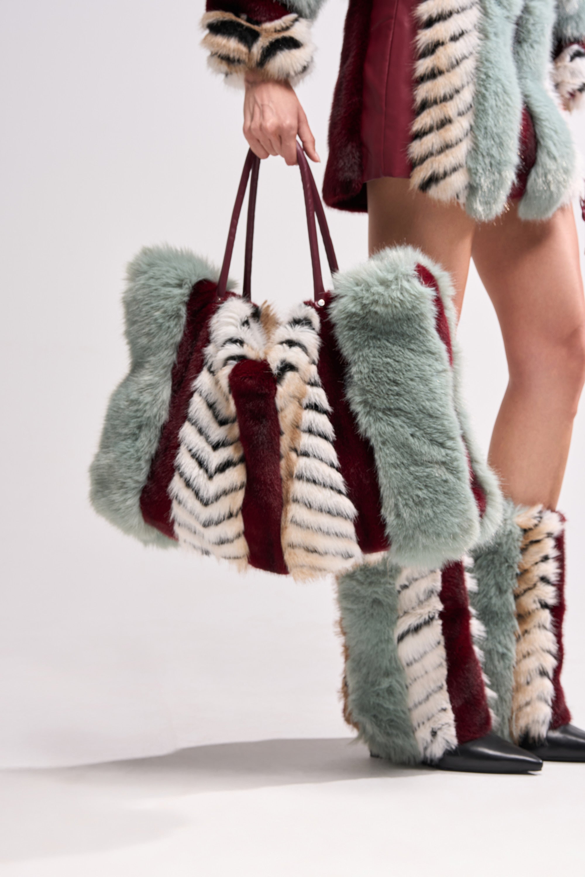 Wearing a green, burgundy, white, and beige striped faux fur outfit, this person carries the HAVE IT ALL FAUX FUR TOTE. Their skirt and boots match with the same bold stripe pattern for a coordinated, eye-catching style. 0