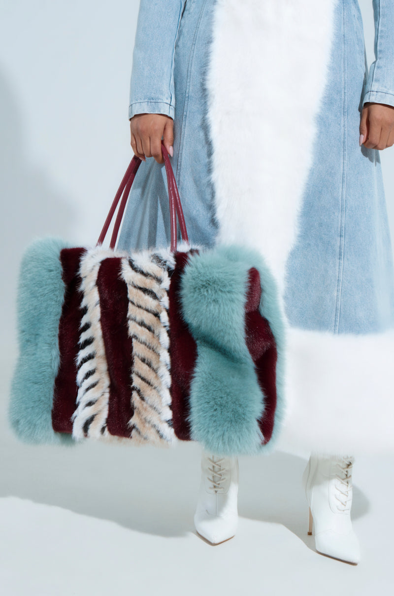 A person in a denim and white fur coat carries the HAVE IT ALL FAUX FUR TOTE, featuring oversized fluff with burgundy, teal, and tan zebra stripes. Chic white heeled boots complete the look, perfectly complementing this statement accessory. 5