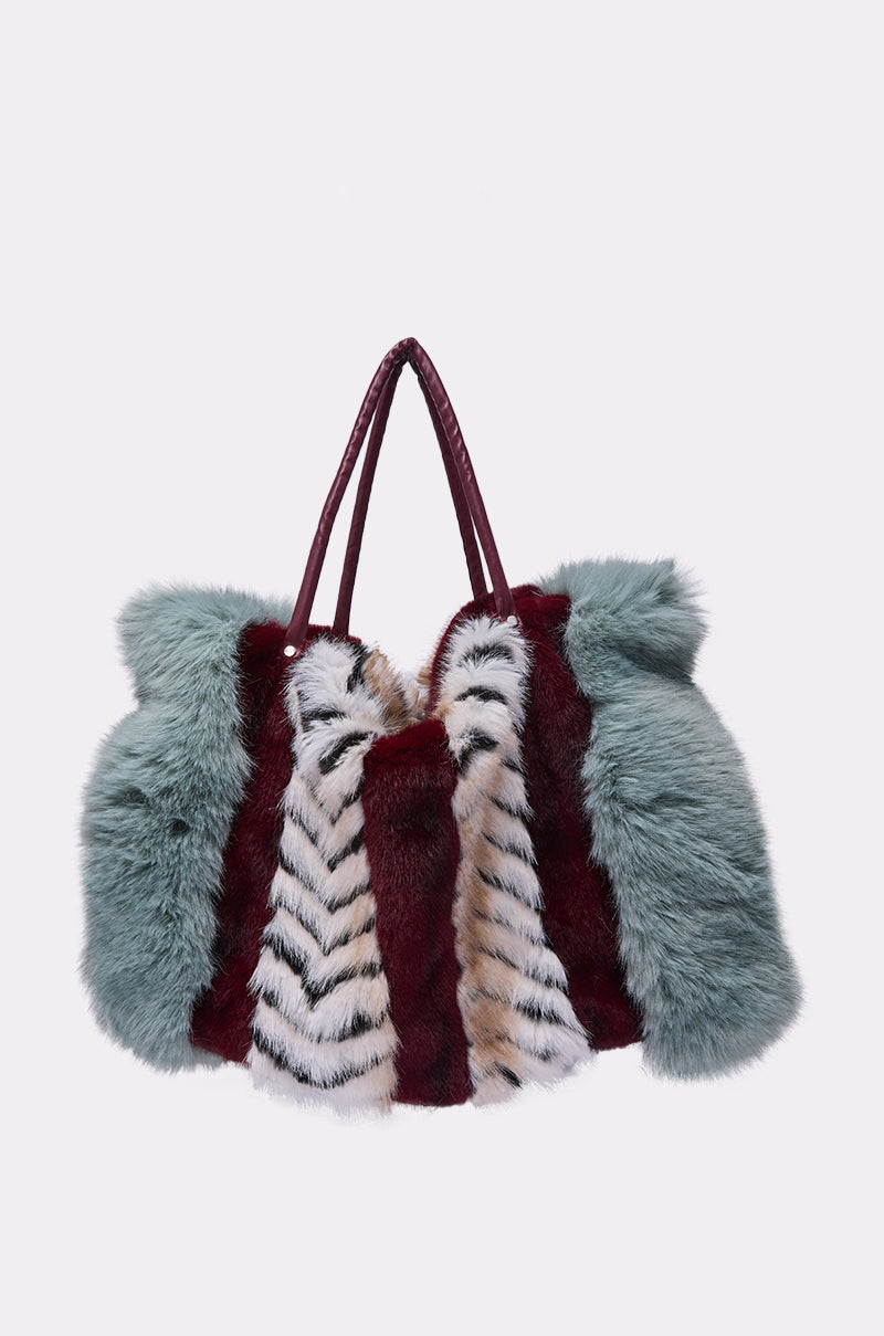 The HAVE IT ALL FAUX FUR TOTE is a large, stylish color block handbag with maroon handles and panels of fluffy faux fur in teal, burgundy, and white with black stripes, shown against a plain light gray background.