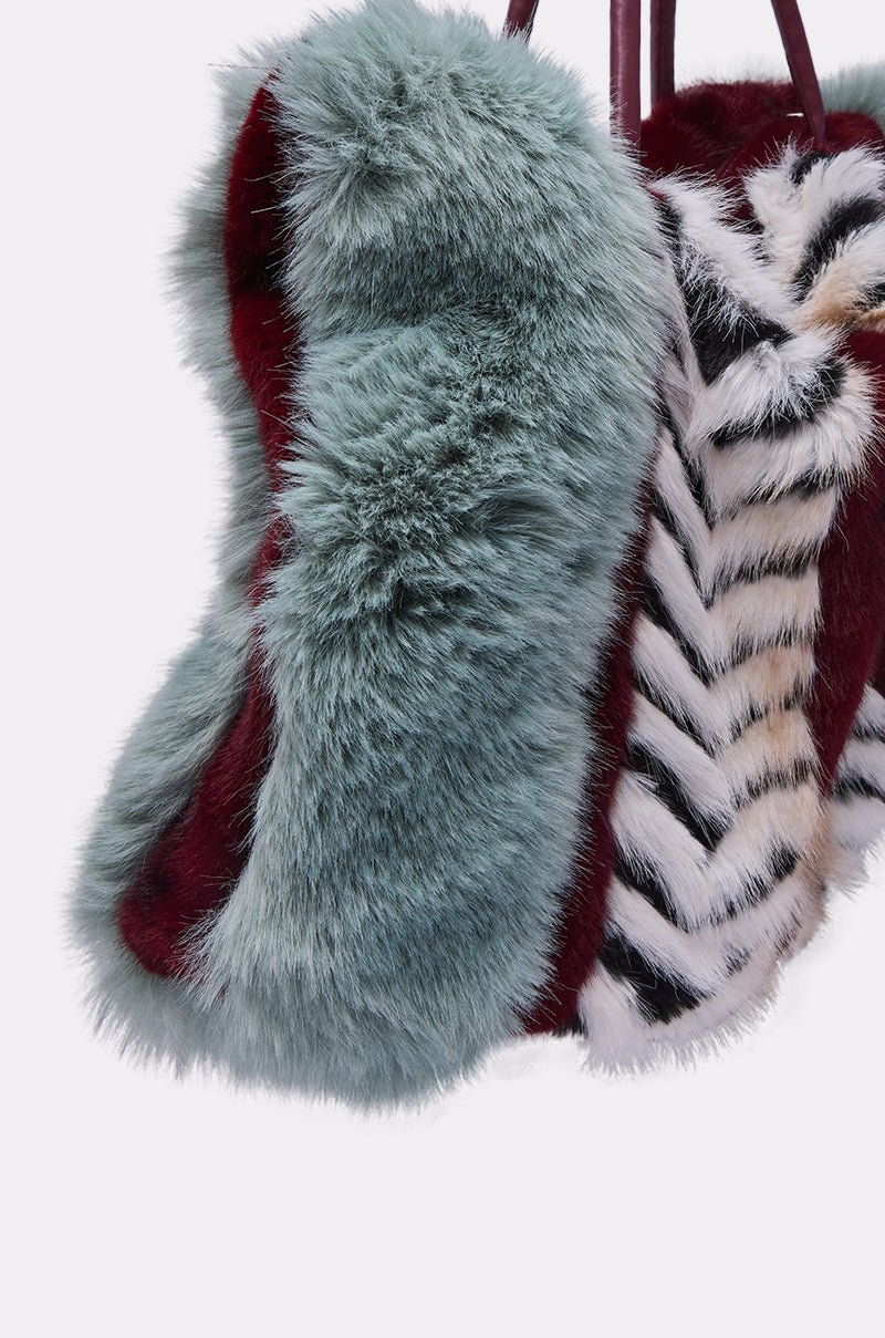 A close-up of the HAVE IT ALL FAUX FUR TOTE, an oversized handbag made from soft faux fur in teal, burgundy, black, and white with a striped chevron pattern and solid color panels.