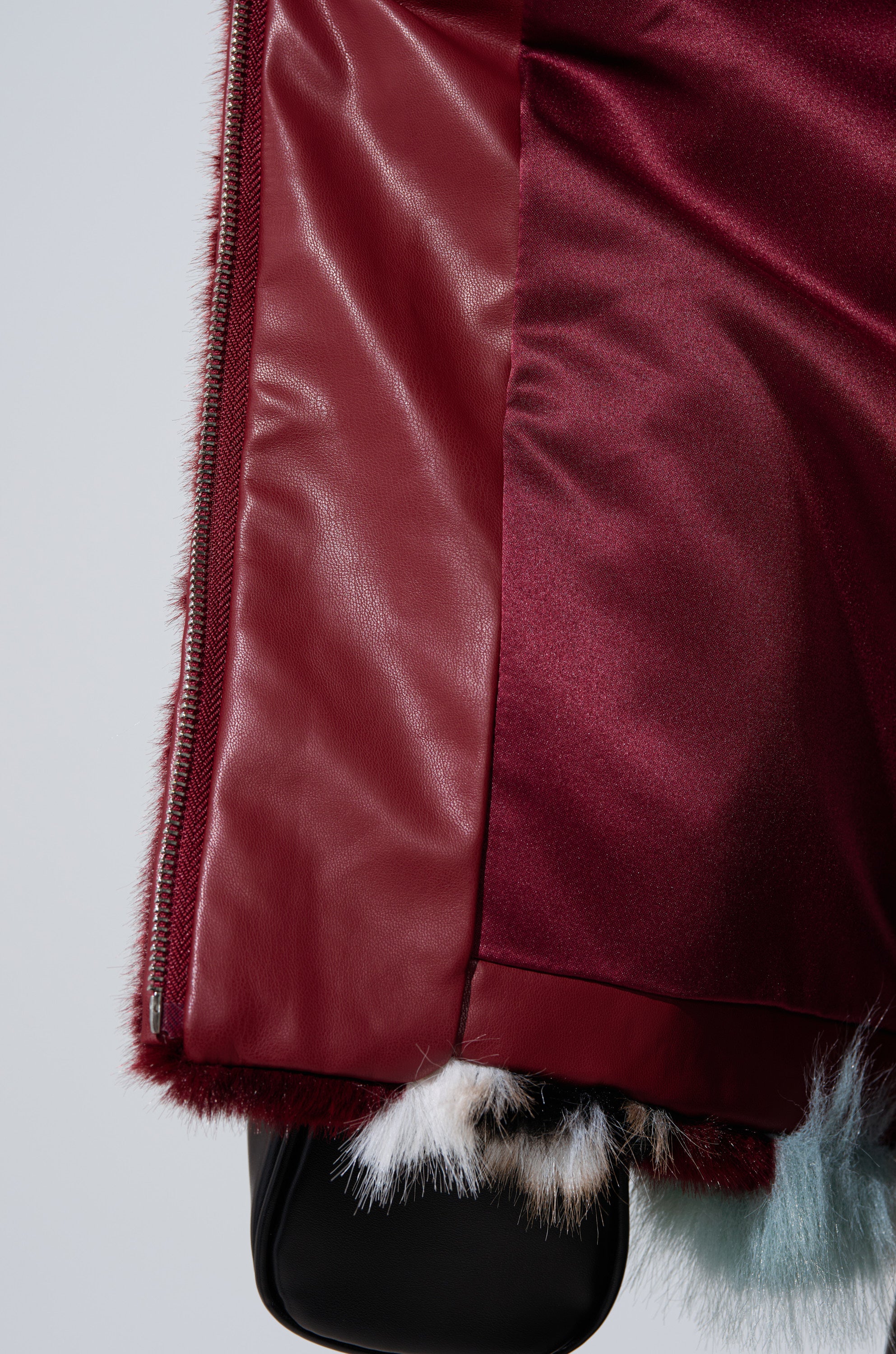 Close-up of the HAVE IT ALL PANELED FUR COAT by AKIRA, featuring burgundy leather, satin-like fabric, a zipper, and white, black & light blue multi-colored faux fur trim at the hem—ideal for bold faux fur coat enthusiasts.