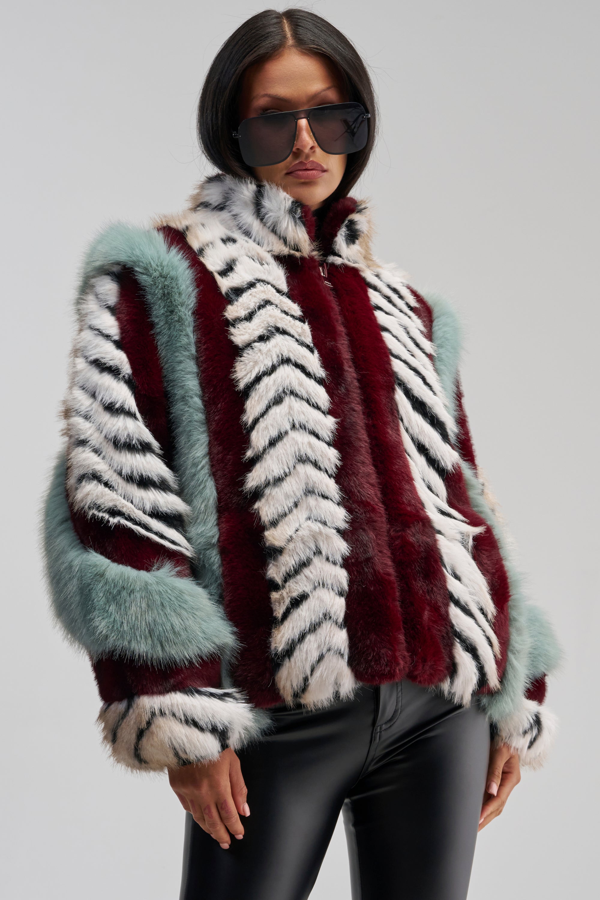 A person wearing large black sunglasses and the HAVE IT ALL PANELED FUR COAT—a multicolored faux fur jacket featuring red, white, black, and teal stripes in an oversized silhouette—paired with shiny black pants stands against a plain, light gray background.