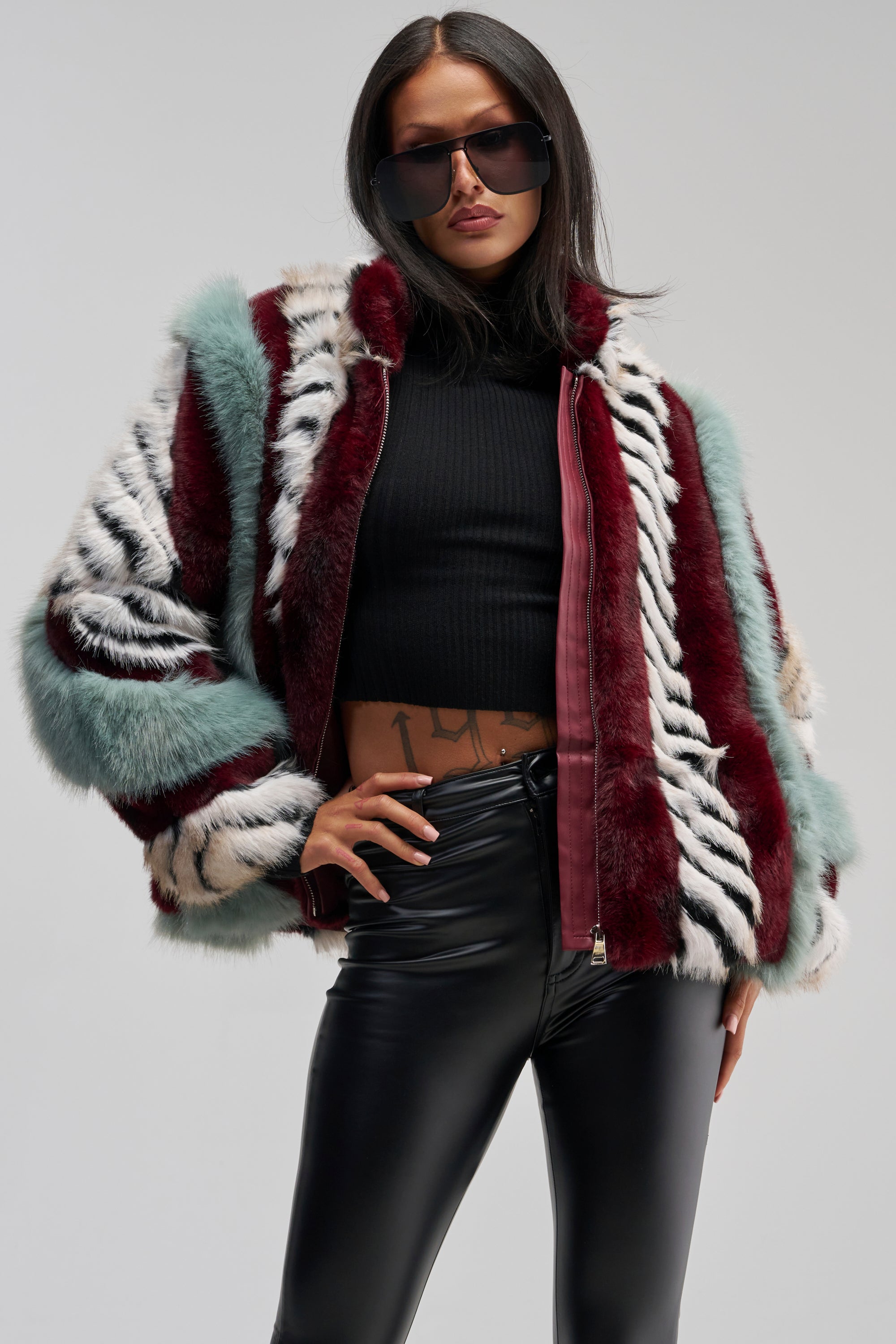 A woman in large sunglasses wears the HAVE IT ALL PANELED FUR COAT—a multicolored faux fur jacket with zebra stripes—styled with a black crop top and black leather pants, standing confidently with one hand on her hip.