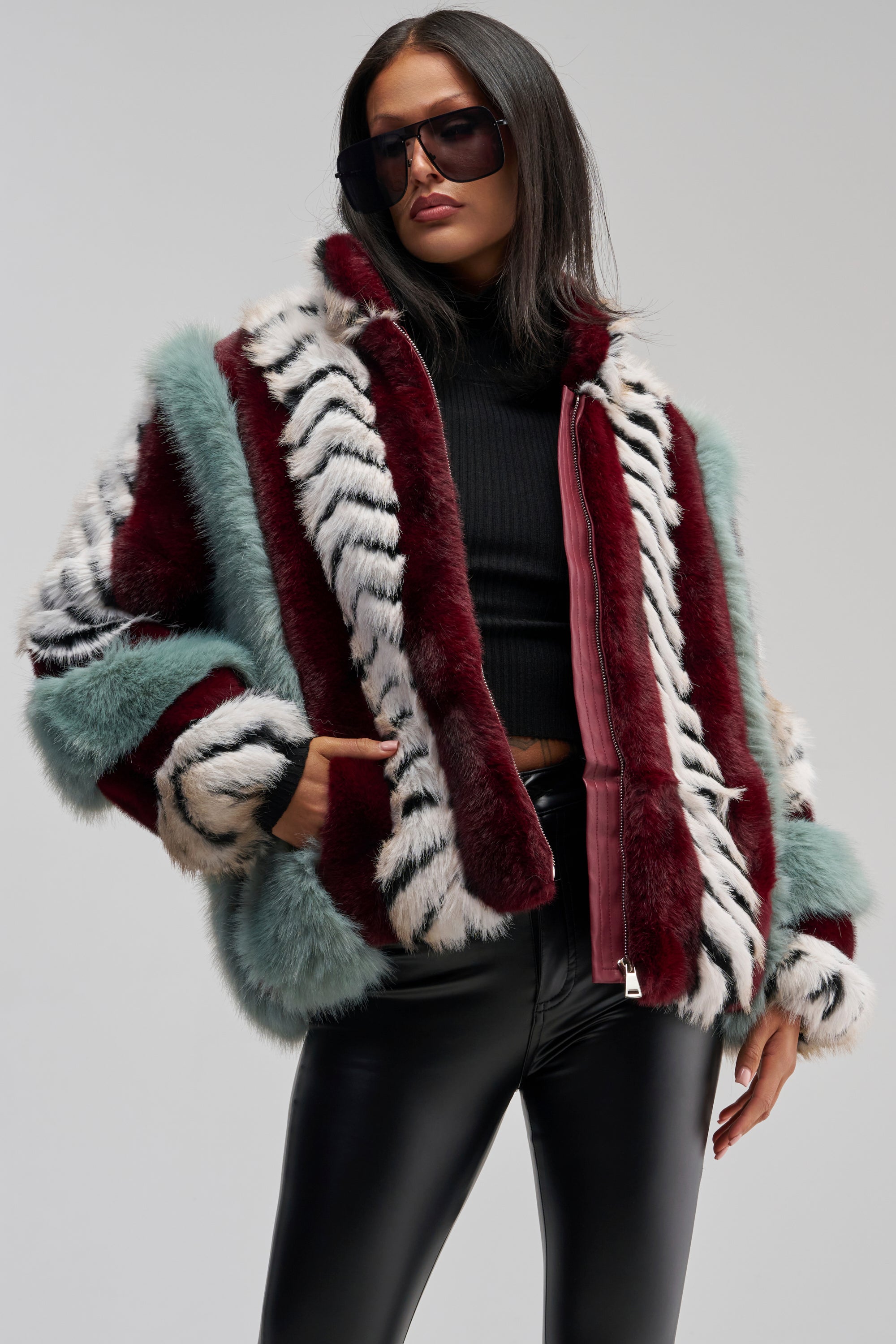A woman poses against a neutral background wearing the HAVE IT ALL PANELED FUR COAT—a colorful faux fur jacket with an oversized silhouette and red, white, and teal paneled patterns—styled with a black turtleneck, black leather pants, large sunglasses, and one hand in her pocket.