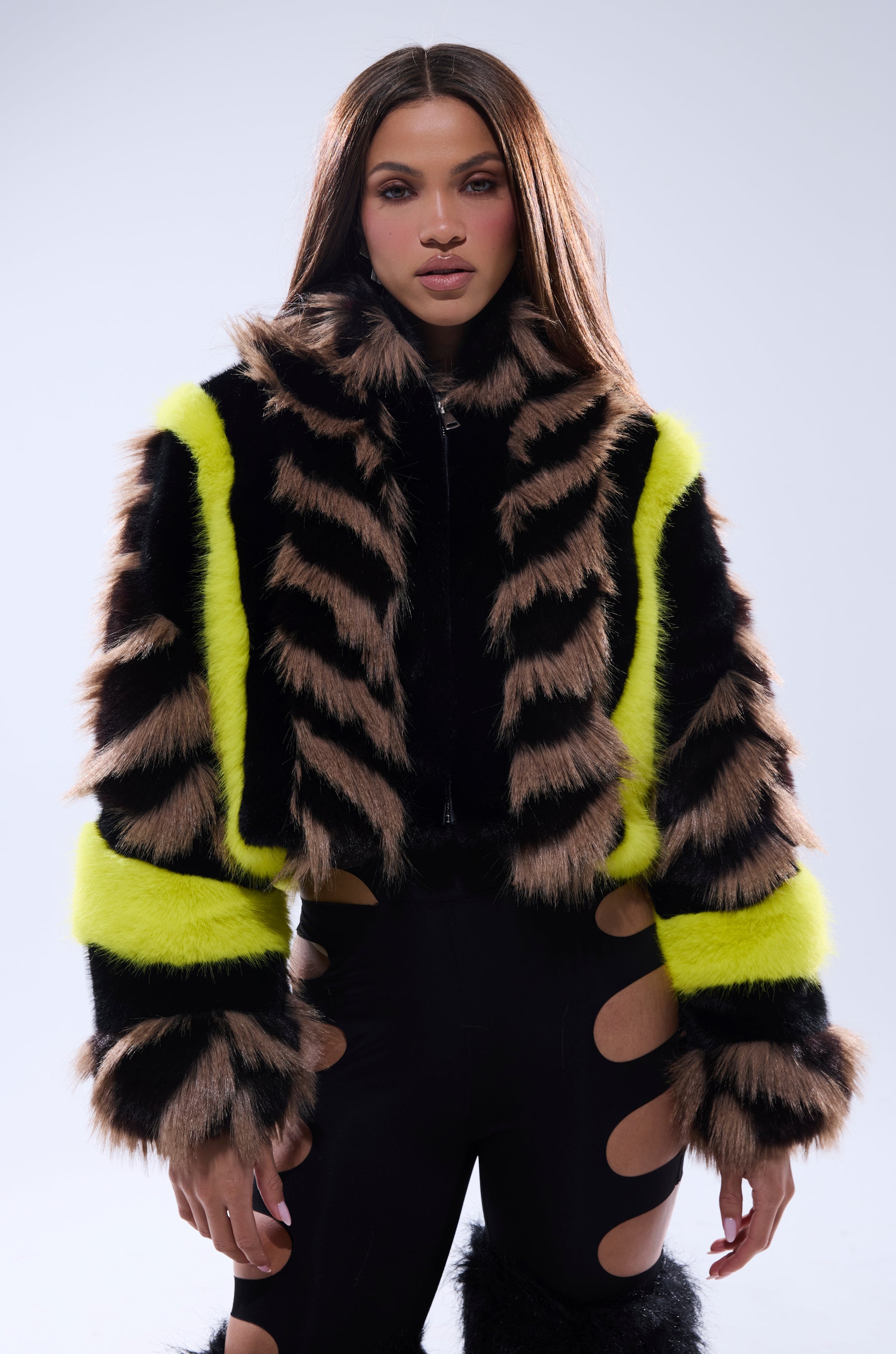 A woman with long, straight brown hair models the MISS ME WITH THAT PANELED FAUX FUR jacket—black, brown, and neon yellow striped—paired with black pants featuring bold side cutouts, set against a plain light gray background.