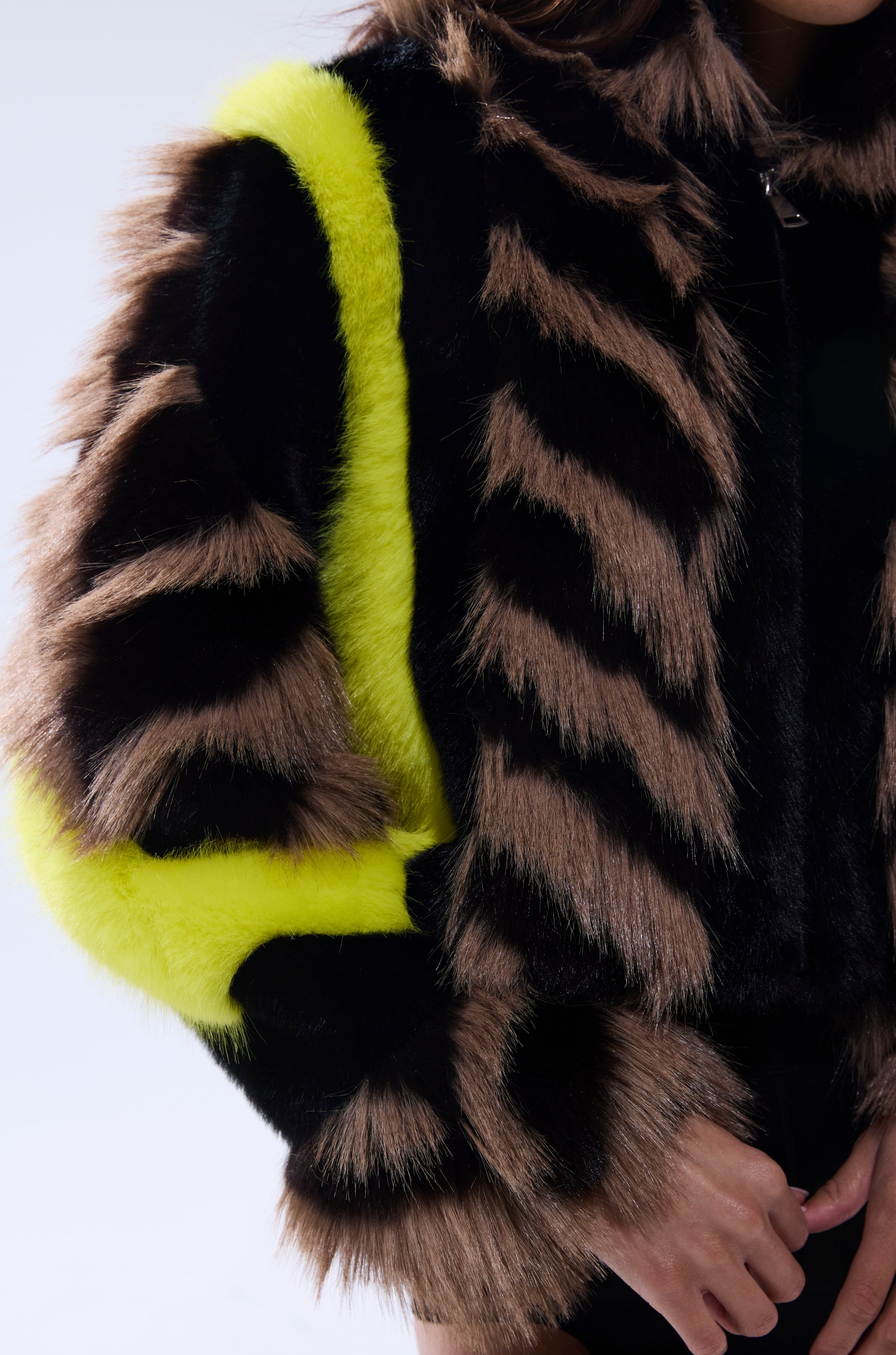 A close-up of a person wearing the MISS ME WITH THAT PANELED FAUX FUR jacket, featuring diagonal black and brown stripes and a bold yellow fur trim on the sleeve. The soft, fluffy jacket is shown without the person's face visible.