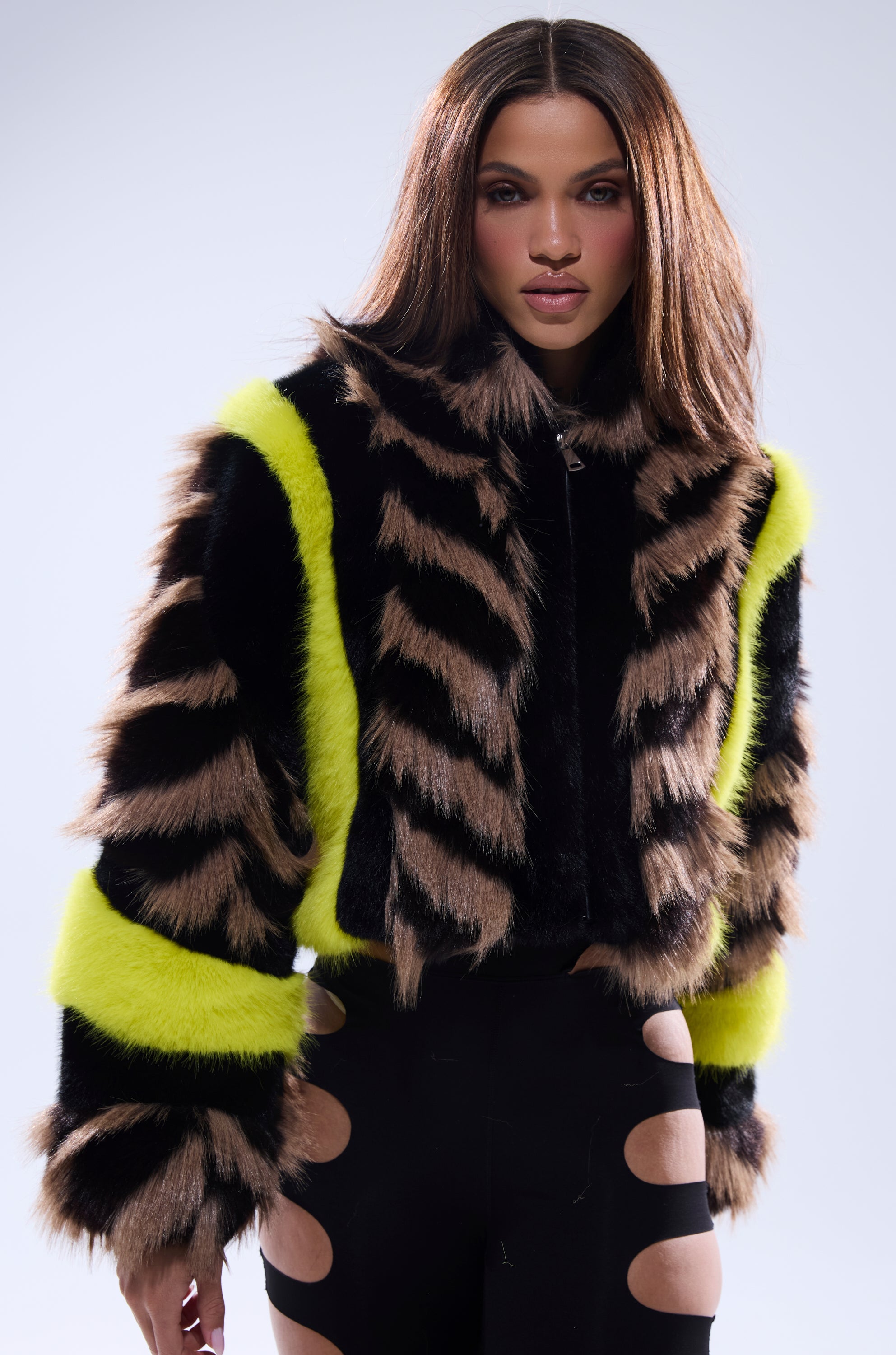 A woman with long brown hair models the MISS ME WITH THAT PANELED FAUX FUR jacket, featuring black and tan tiger stripes with yellow trim, paired with black pants with large oval leg cutouts, standing against a plain background.