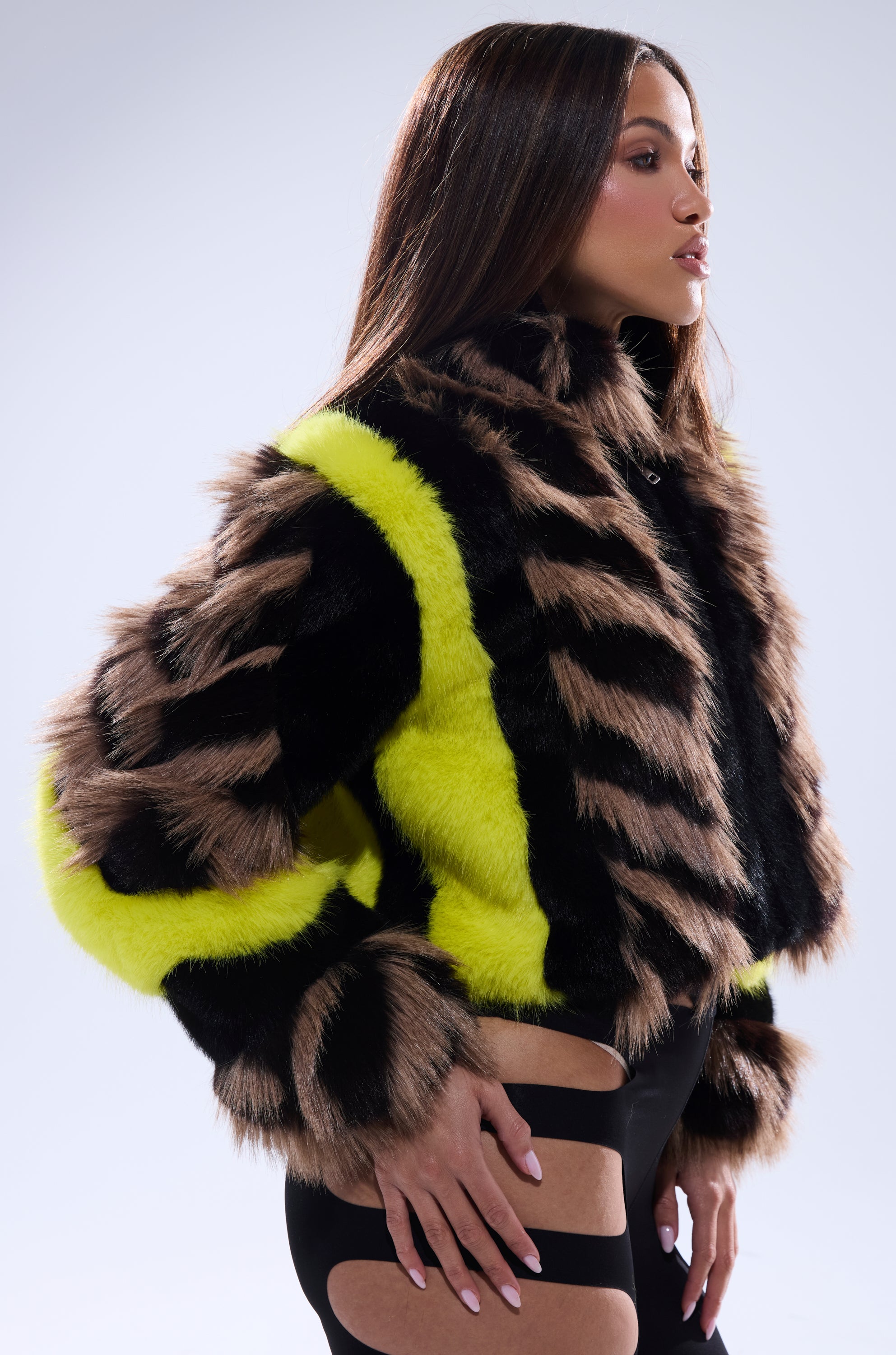 A woman poses in the "MISS ME WITH THAT PANELED FAUX FUR" jacket, featuring cropped, paneled faux fur with brown, black, and bright yellow stripes, paired with black cutout pants against a plain light background.