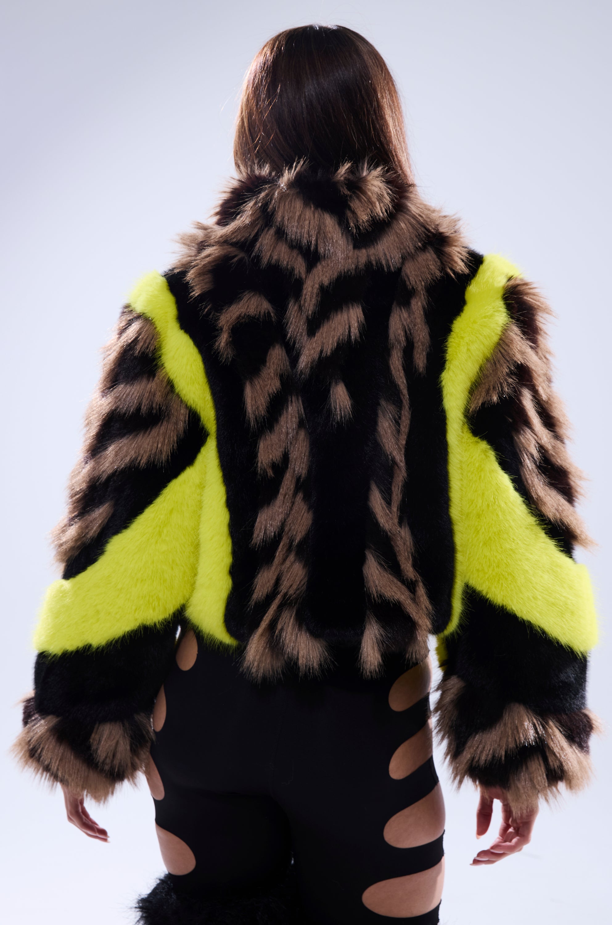 A person with long brown hair, seen from behind, wears the MISS ME WITH THAT PANELED FAUX FUR jacket with black, brown, and neon yellow patterns, paired with black pants that have large oval cutouts along the legs.