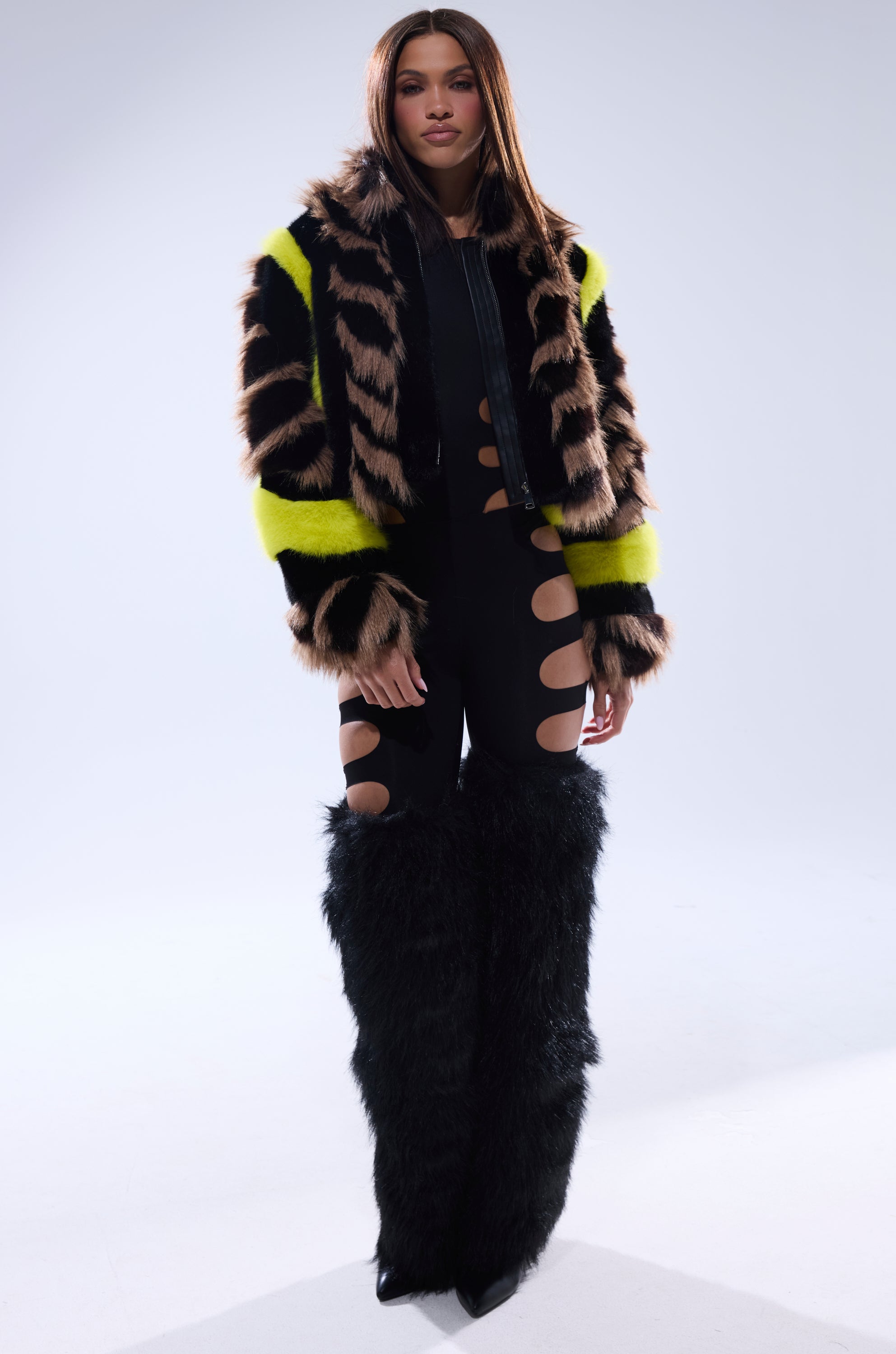 A woman models the MISS ME WITH THAT PANELED FAUX FUR jacket with black, brown, and neon yellow stripes, paired with black cut-out leggings and tall black furry leg warmers against a simple background that highlights her style.
