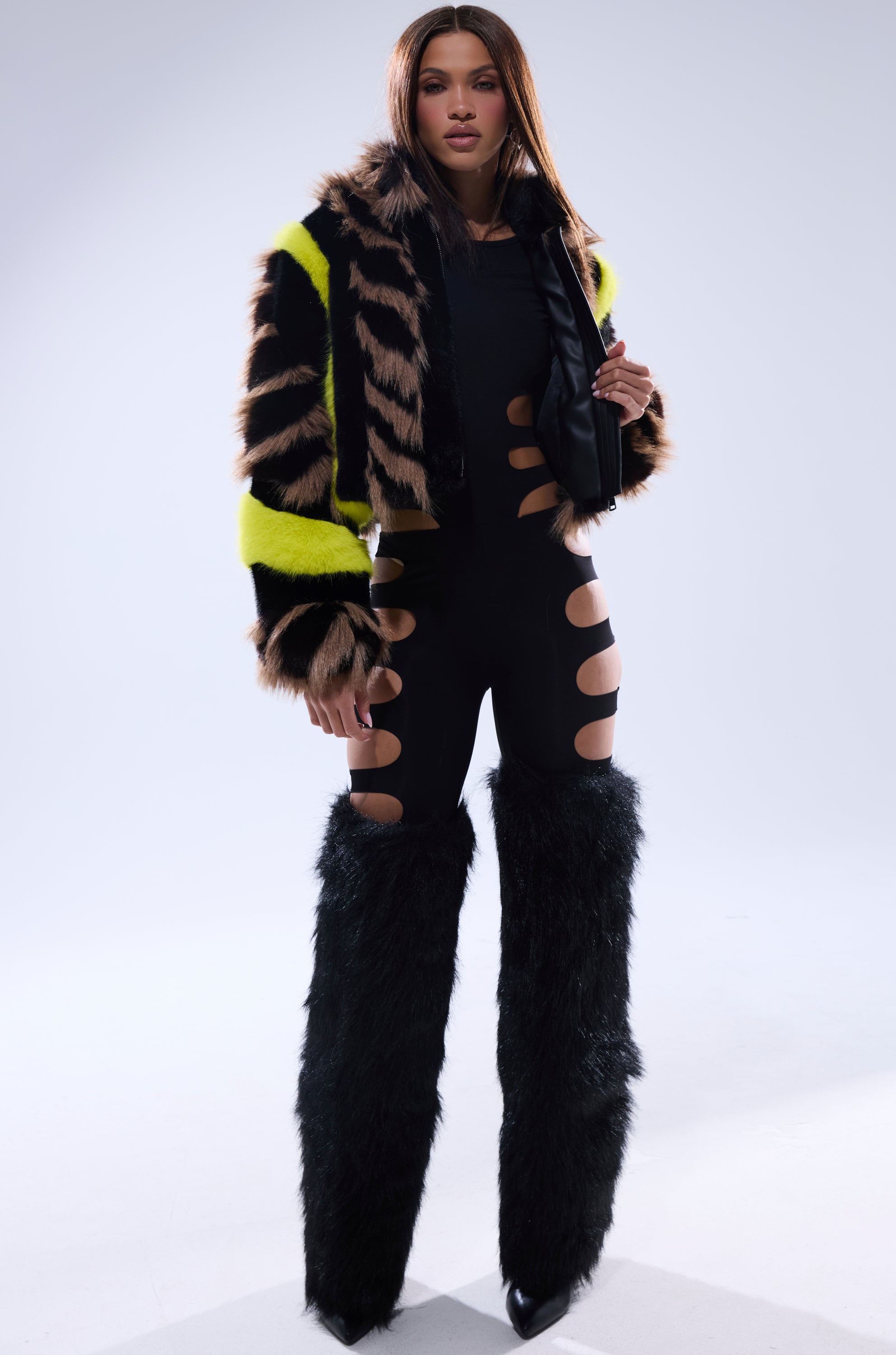 A woman stands confidently in the MISS ME WITH THAT PANELED FAUX FUR jacket with yellow contrast trim, a black cutout bodysuit, and tall black furry chaps over pointed shoes against a plain light background.