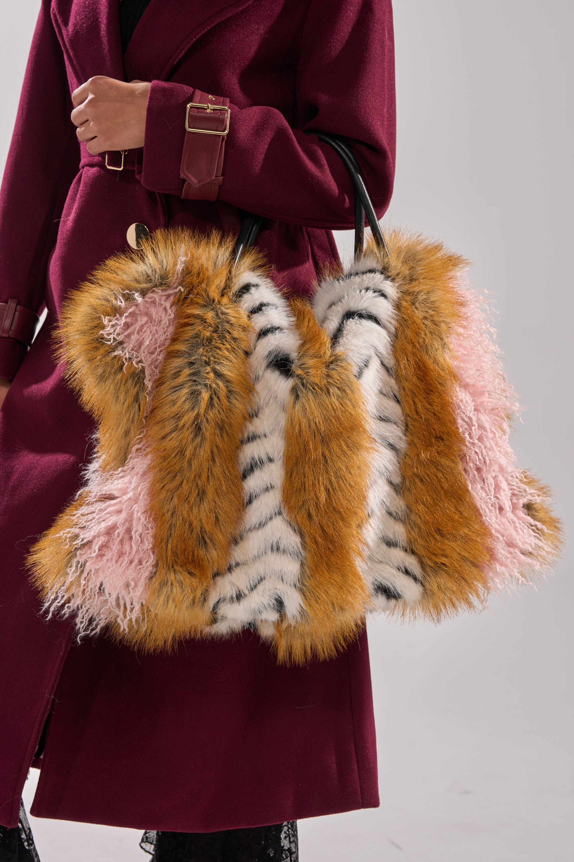 Someone in a burgundy coat carries the WILD ONE FAUX FUR TOTE, an oversized plush bag in orange, pink, and white with bold black stripes and sleek black handles, designed to resemble animal fur patterns. 1