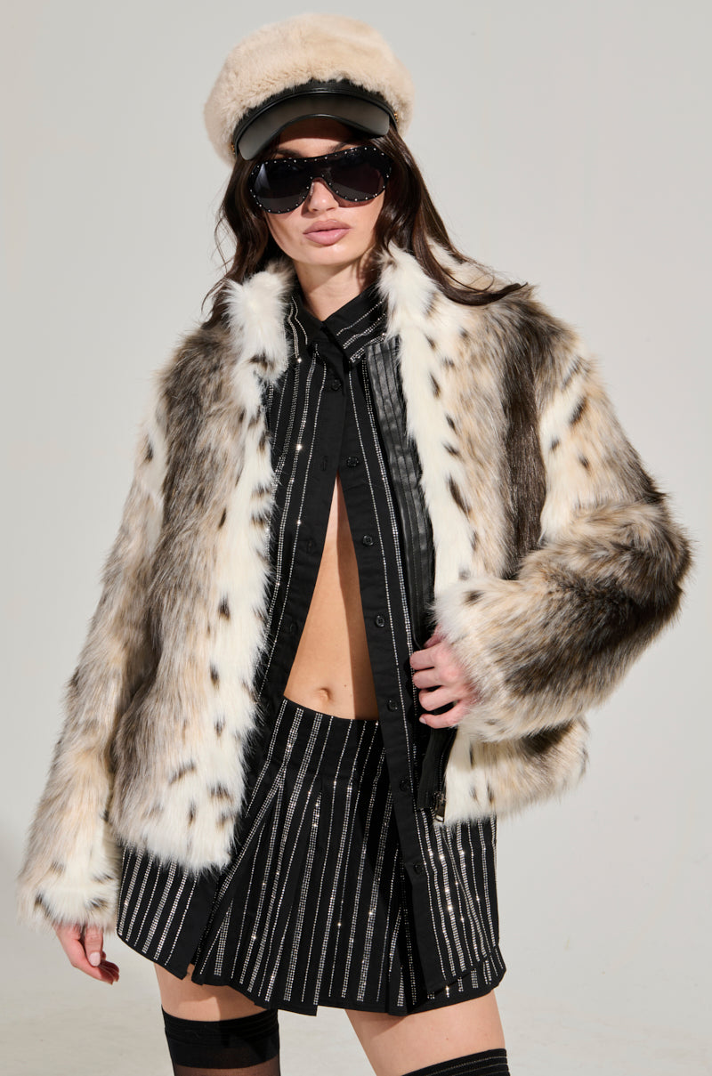 A person wearing the ELSIE FAUX FUR BOMBER, stylish sunglasses, and a cream-colored hat. They are dressed in a black outfit with vertical stripes on both the sleeves and skirt.