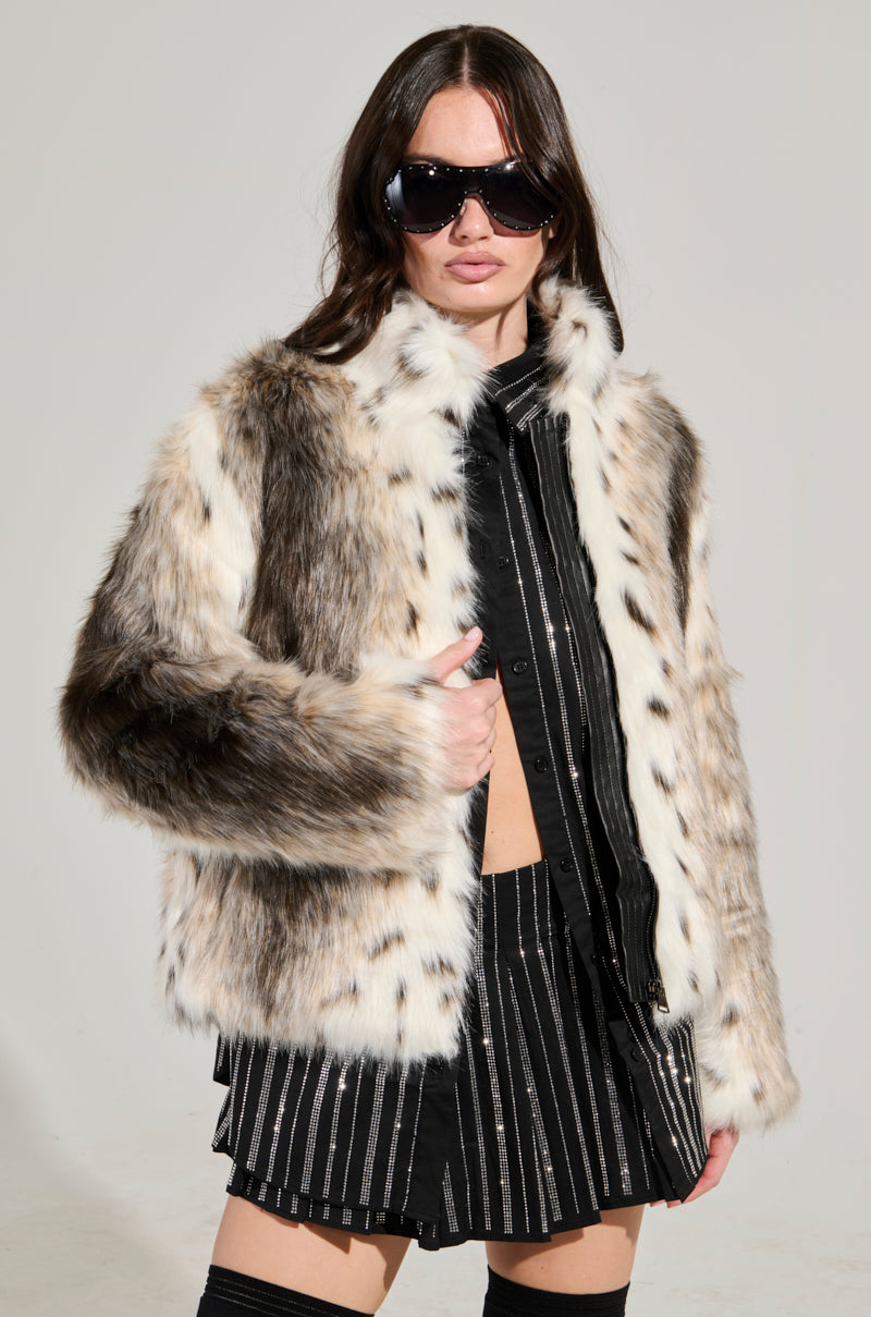 A person wearing oversized sunglasses and the ELSIE FAUX FUR BOMBER, featuring a chic animal print mix of brown, white, and black, poses against a plain background. Their hair is loose, and they exude confidence in their sleek, striped outfit.