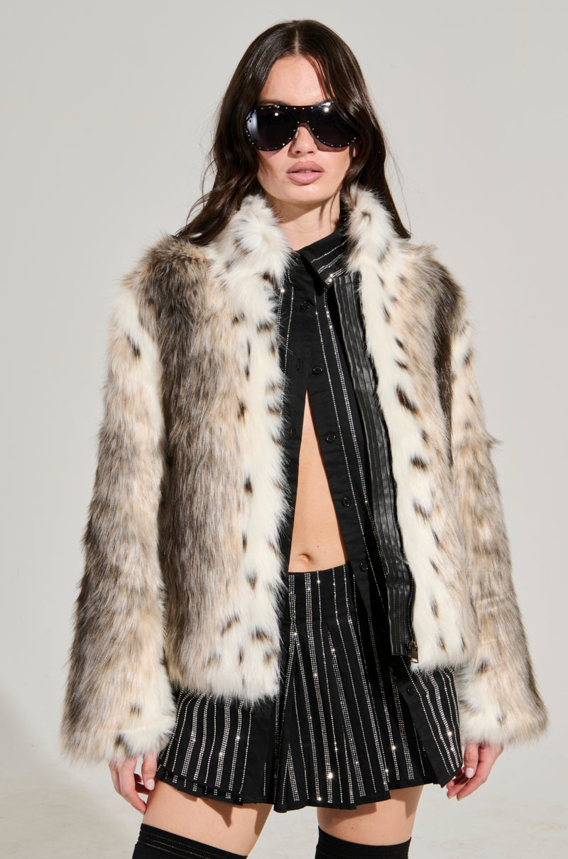 A person wearing the ELSIE FAUX FUR BOMBER, a black shirt, a striped skirt, and large sunglasses stands against a plain background. The jacket showcases a blend of white, brown, and black colors in an animal print style.